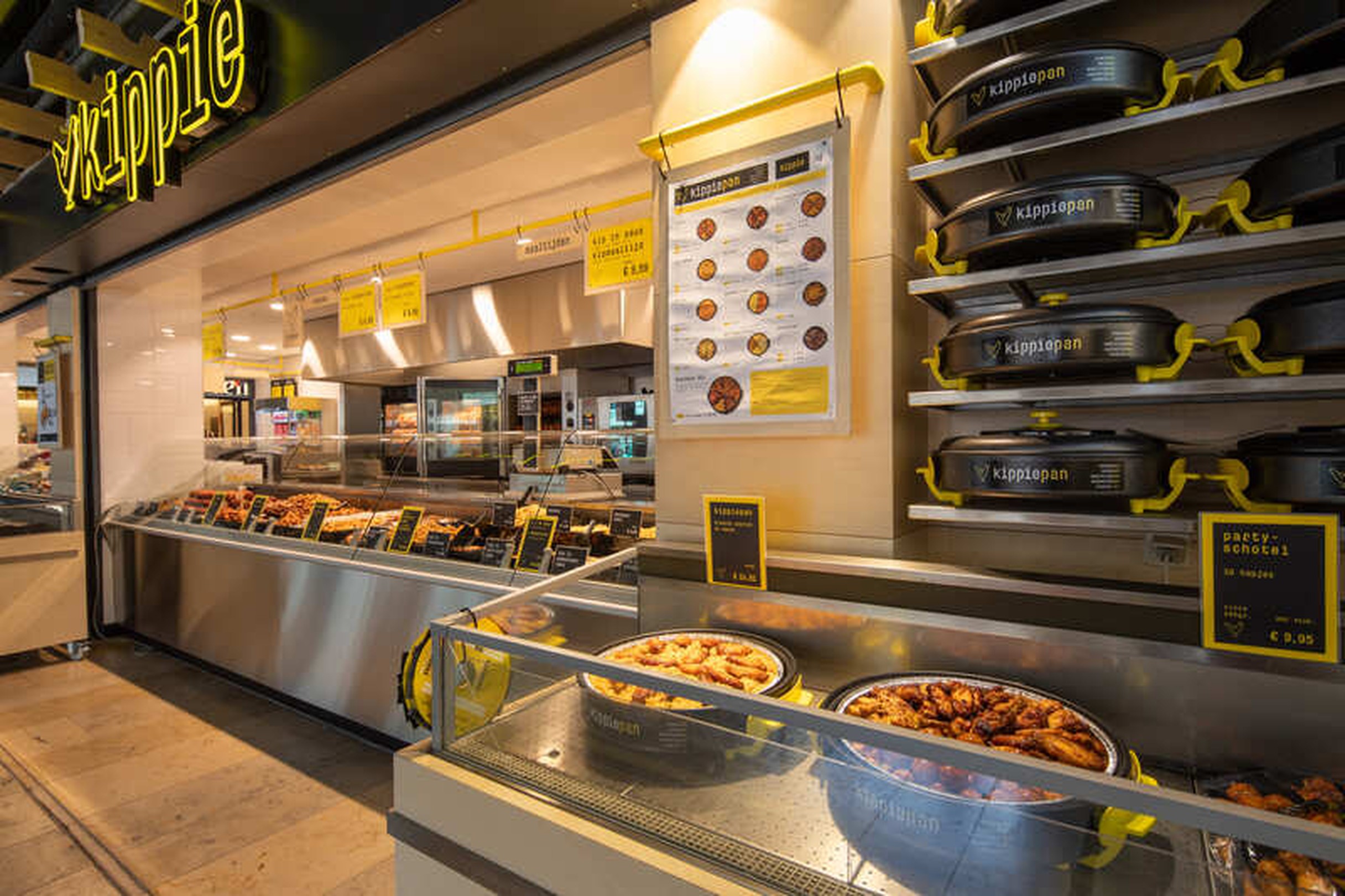 Vooraanzicht van Kippie Nijmegen, een Grill & Maaltijdenwinkel in een winkelcentrum, met glazen toonbanken vol gegrilde gerechten en maaltijden. Aan de gevel hangen meerdere Kippie logo’s en borden.