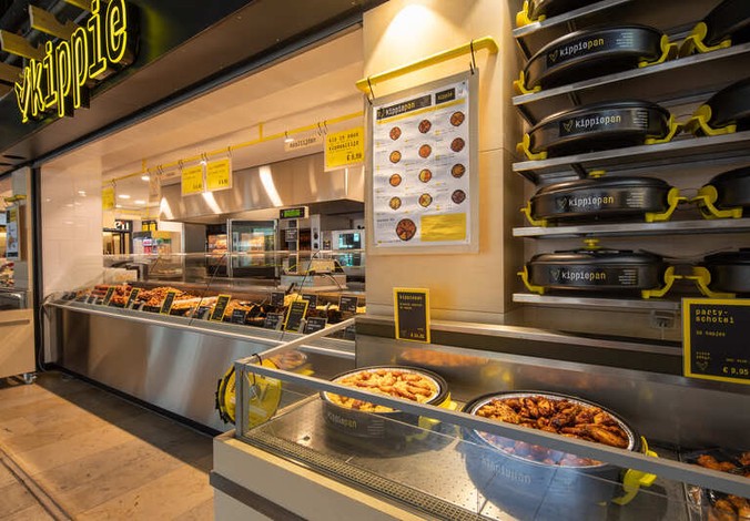 Vooraanzicht van Kippie Nijmegen, een Grill & Maaltijdenwinkel in een winkelcentrum, met glazen toonbanken vol gegrilde gerechten en maaltijden. Aan de gevel hangen meerdere Kippie logo’s en borden.