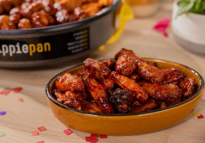 Close-up of a dish filled with glazed chicken wings on a table, with another bowl of mixed appetizers in the background.