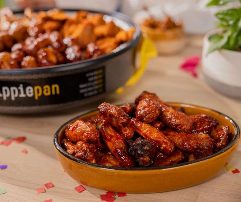 Close-up of a dish filled with glazed chicken wings on a table, with another bowl of mixed appetizers in the background.