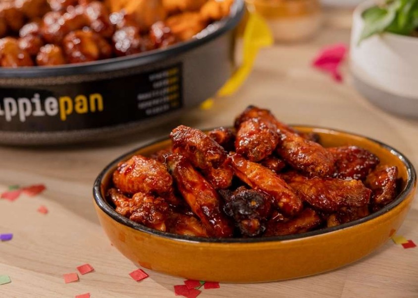 Close-up of a dish filled with glazed chicken wings on a table, with another bowl of mixed appetizers in the background.
