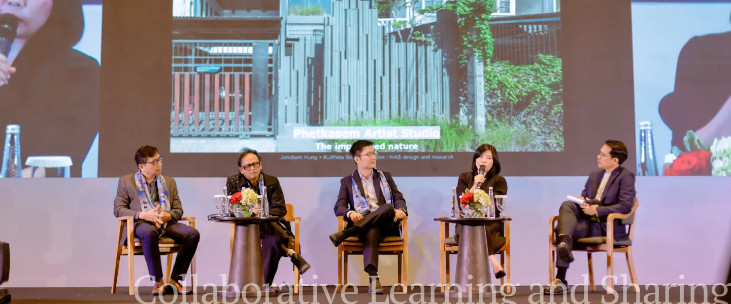 A panel of five speakers sits on a stage discussing, with a large screen behind them displaying an image and text.