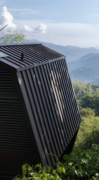A modern, angular metal structure stands amidst lush greenery with a backdrop of misty mountains under a partly cloudy sky.