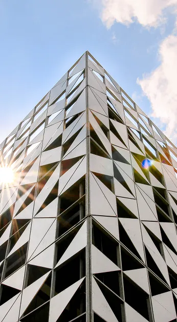 Geometric building with triangular patterns, shot from a low angle against a blue sky, sunburst peeking through.