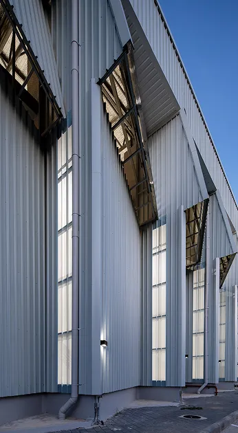 Close-up of a modern industrial building with vertical metal panels and large, angled windows, under a clear blue sky.