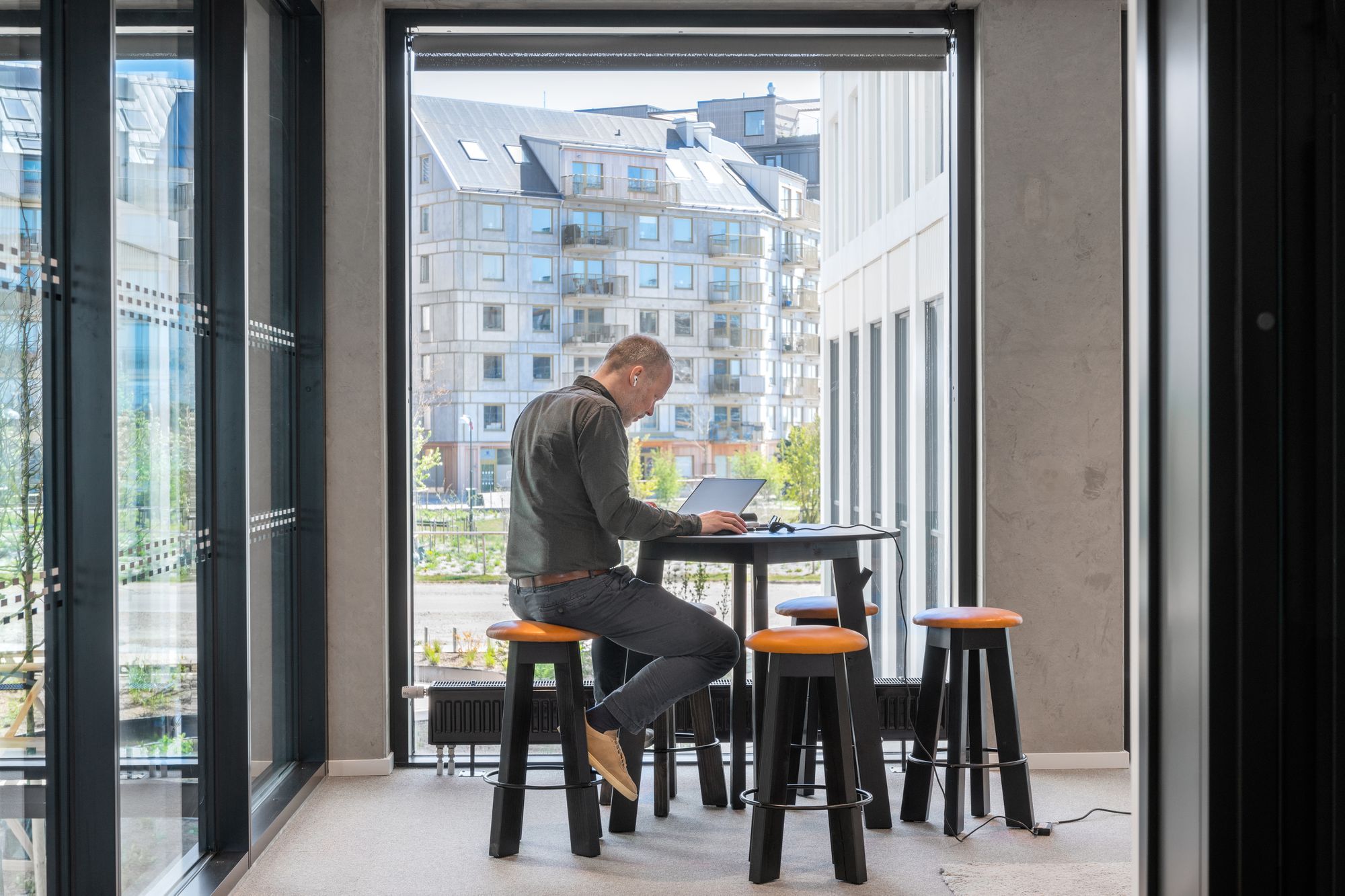Man med hörlurar som jobbar vid en laptop vid ett högt bord intill ett stort fönster med utsikt över bostadshus.