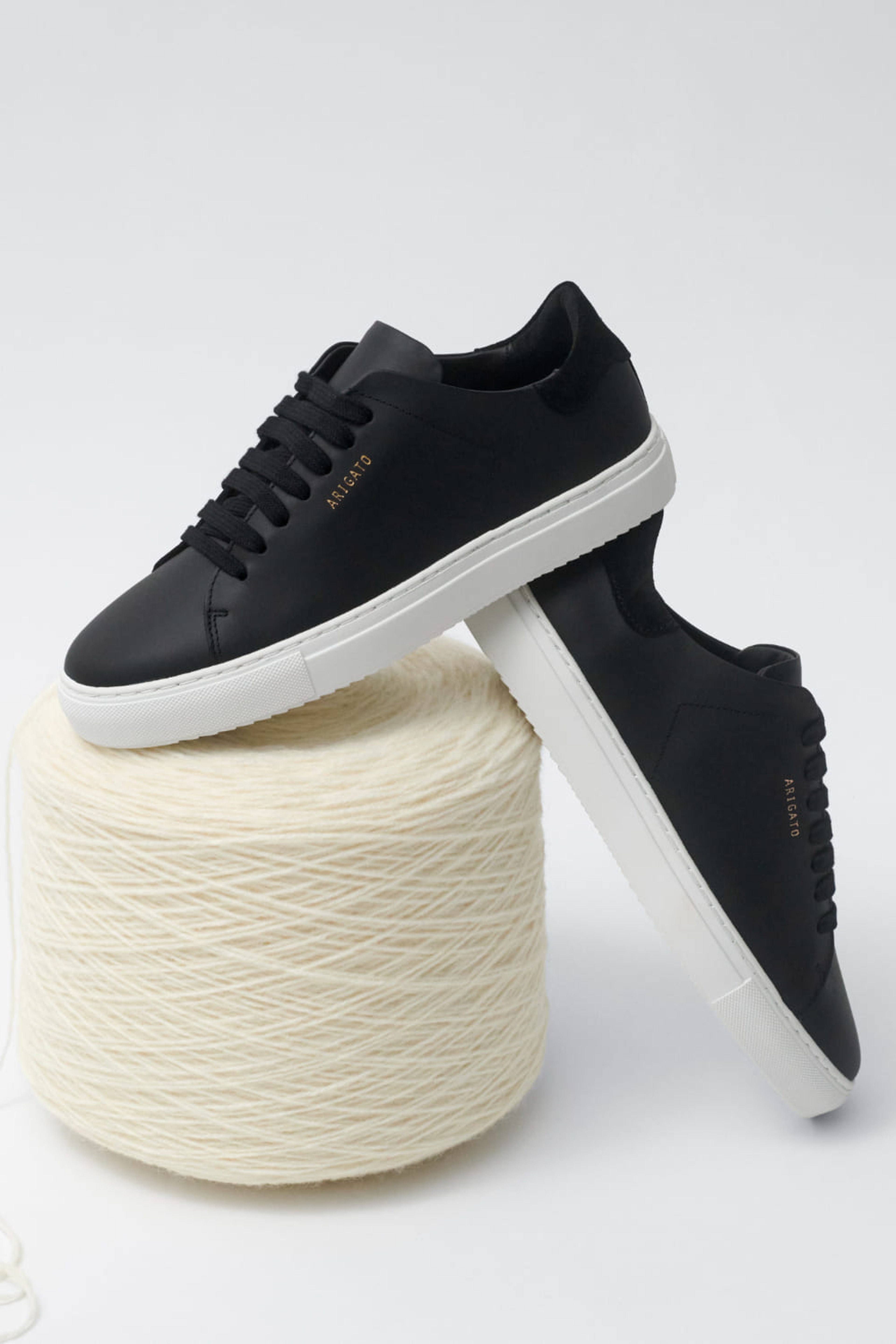 Black sneakers with white soles rest on a large spool of cream yarn, showcasing a casual, stylish design in a minimalist setting.
