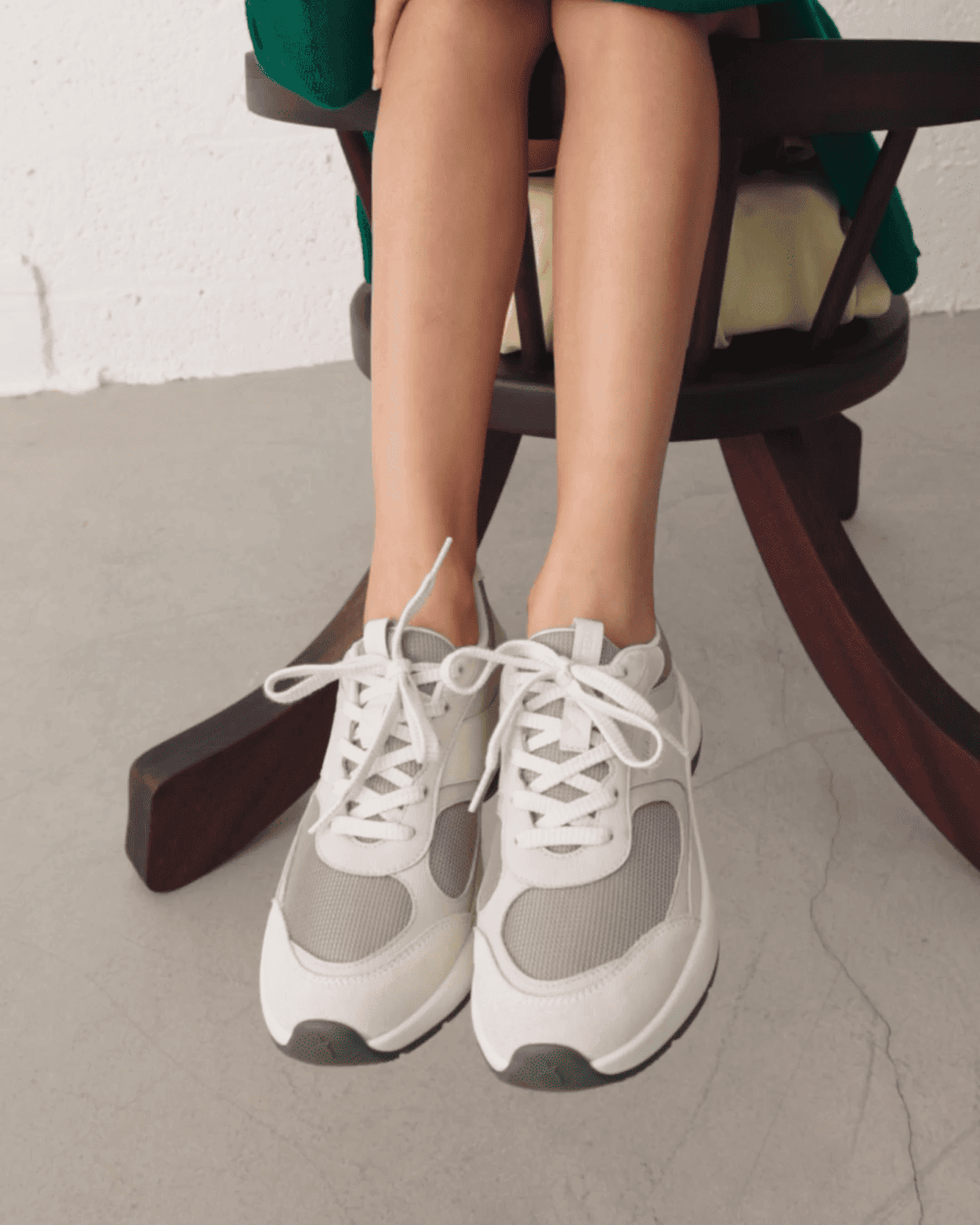 A person sitting on a chair wearing white and gray sneakers with loosely tied laces.