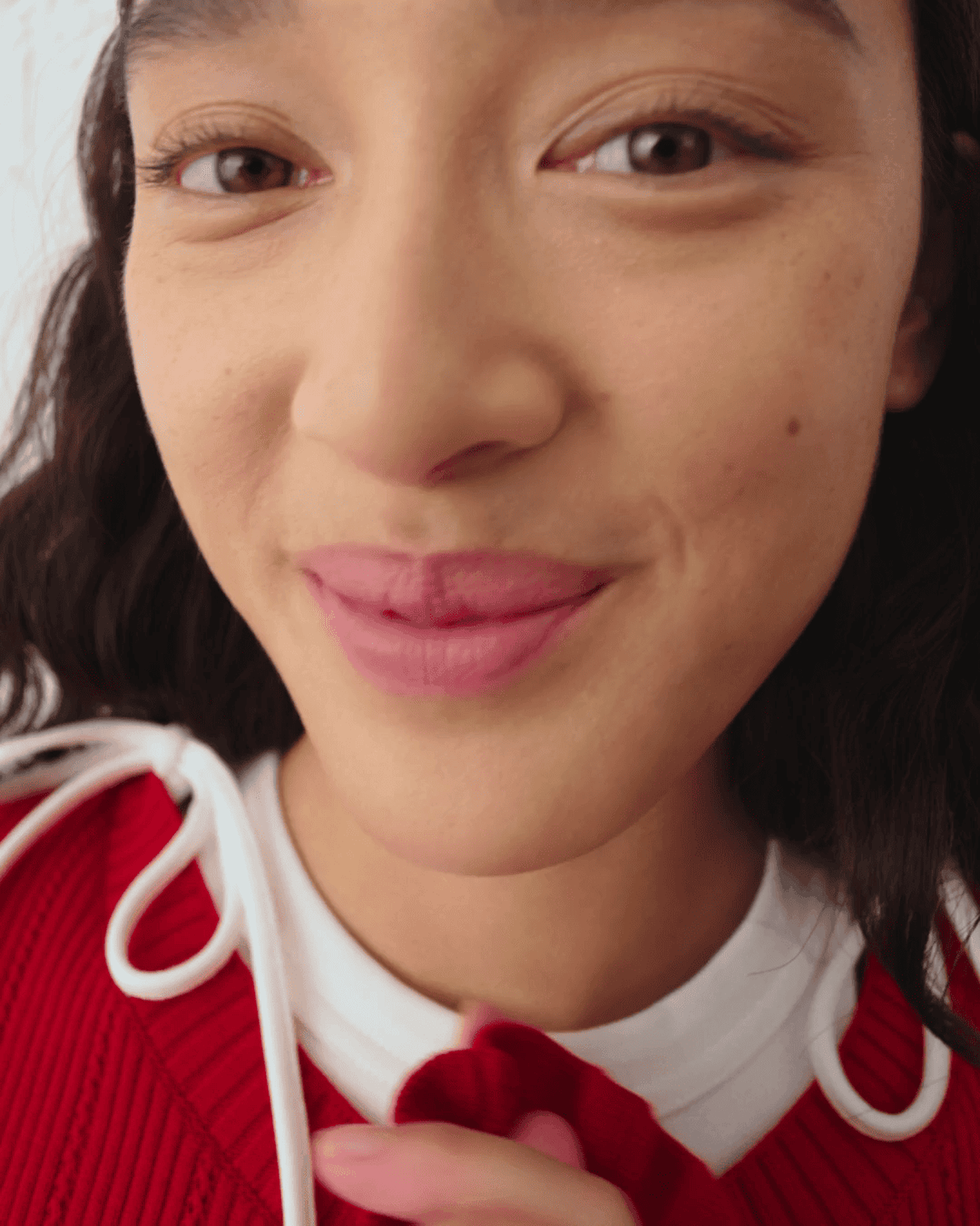 A person smiling and slightly puckering lips, wearing a red sweater with white ties and a white shirt underneath. Close-up view.