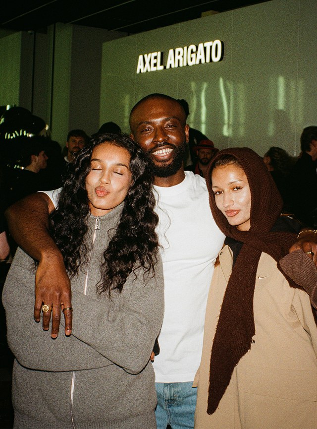 Three people pose happily together; one in a gray jacket, another in white, and the third in a light brown coat with a headscarf.
