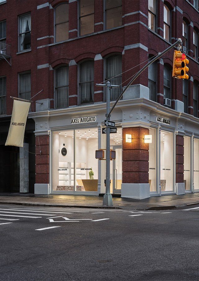 Street corner with a brick building housing the Axel Arigato store, displaying modern interiors through large windows. Traffic light and crosswalk visible.