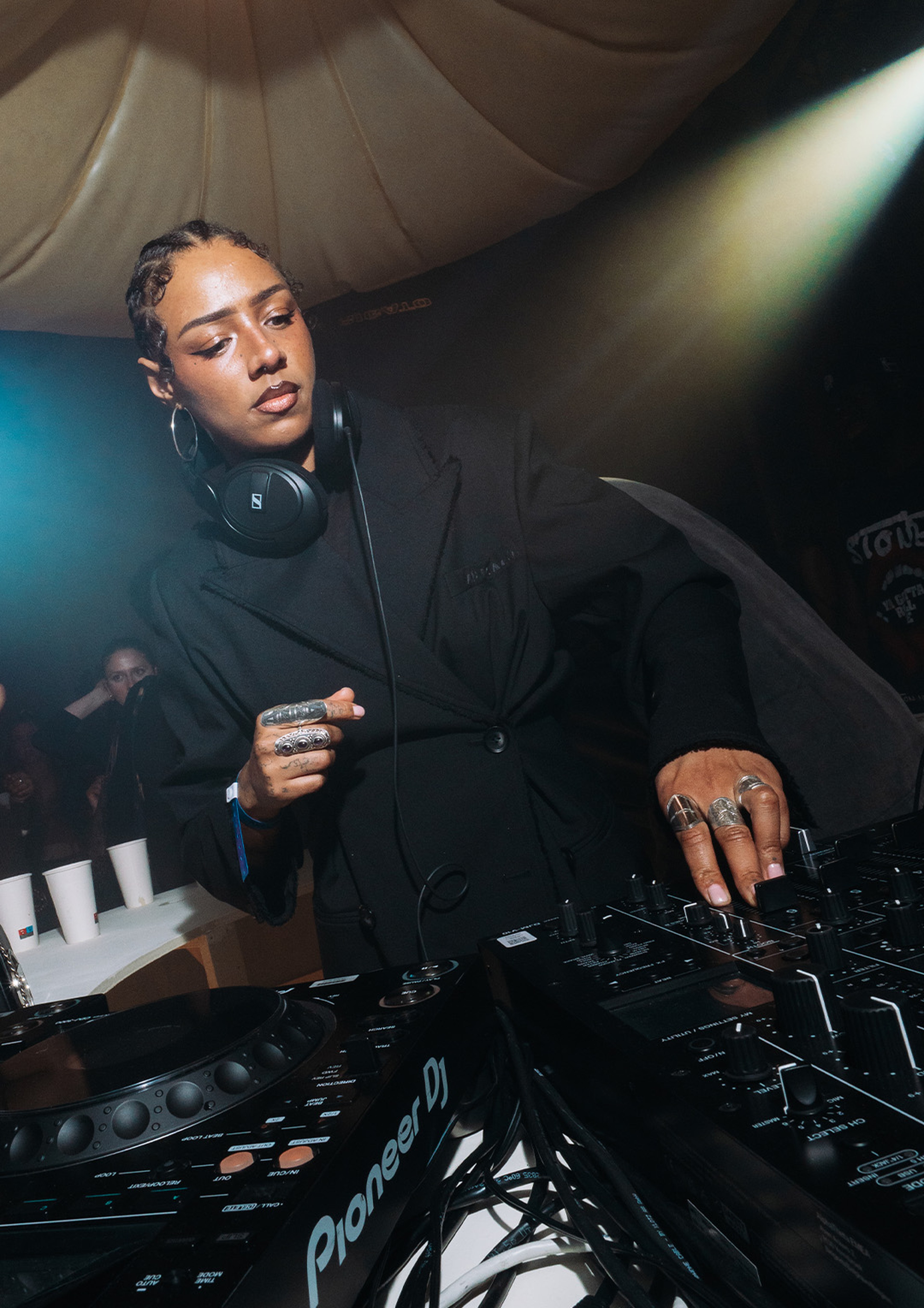 DJ wearing headphones, adjusting the mixer at a booth, focused under stage lighting, with people and cups visible in the dim background.