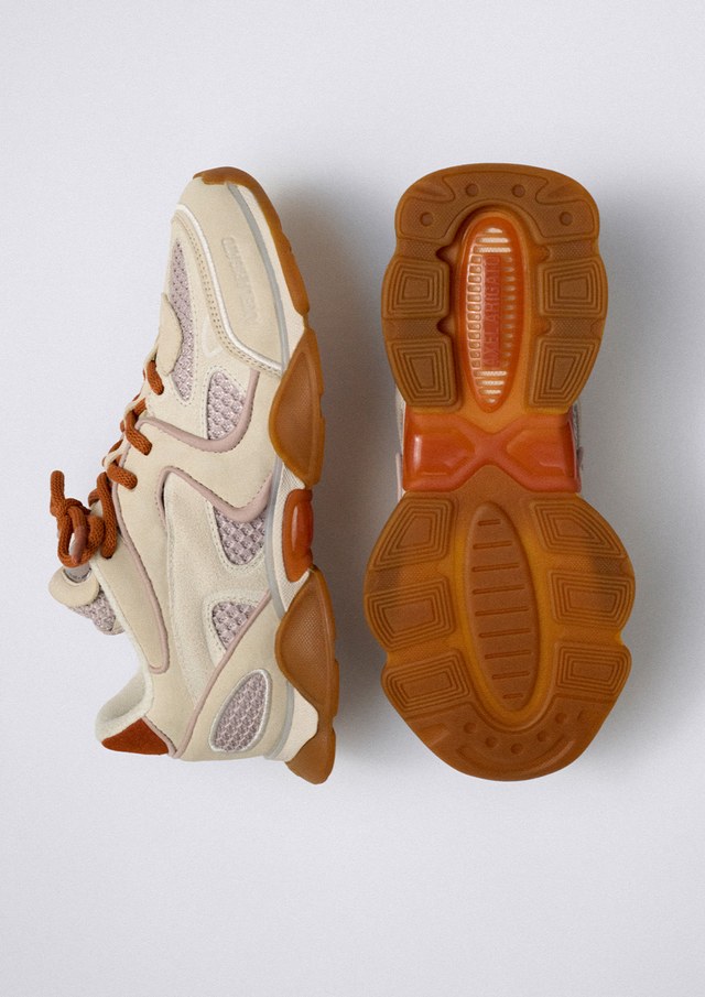 A pair of beige and orange sneakers, with one showing the side profile and the other displaying the gum sole, on a light gray background.