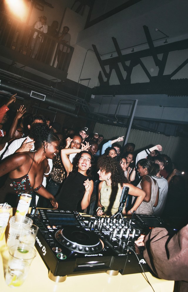 Crowded party scene with people dancing and smiling, DJ equipment in the foreground, and drink cups on a table.