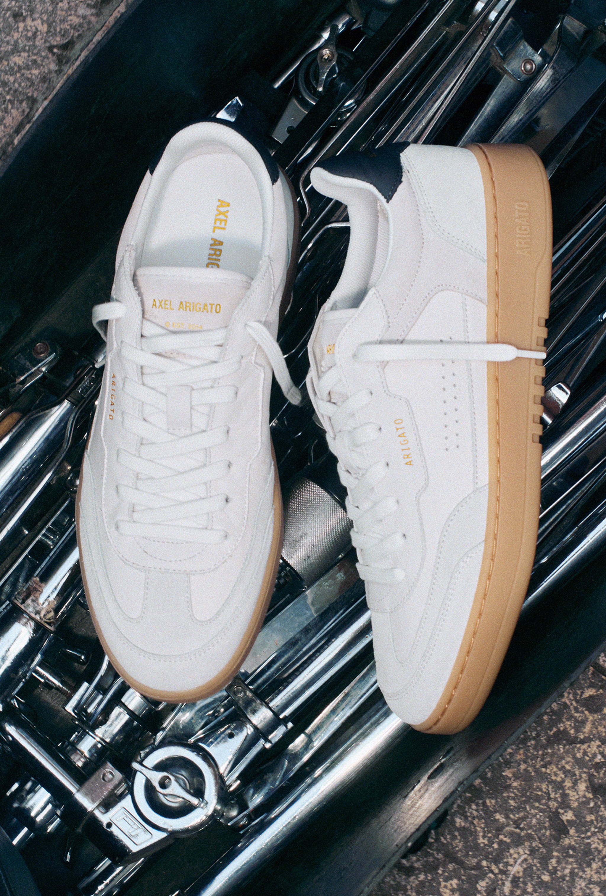 White Axel Arigato sneakers with gum soles placed on a metallic surface, showcasing their sleek design and branding on the tongue and side.