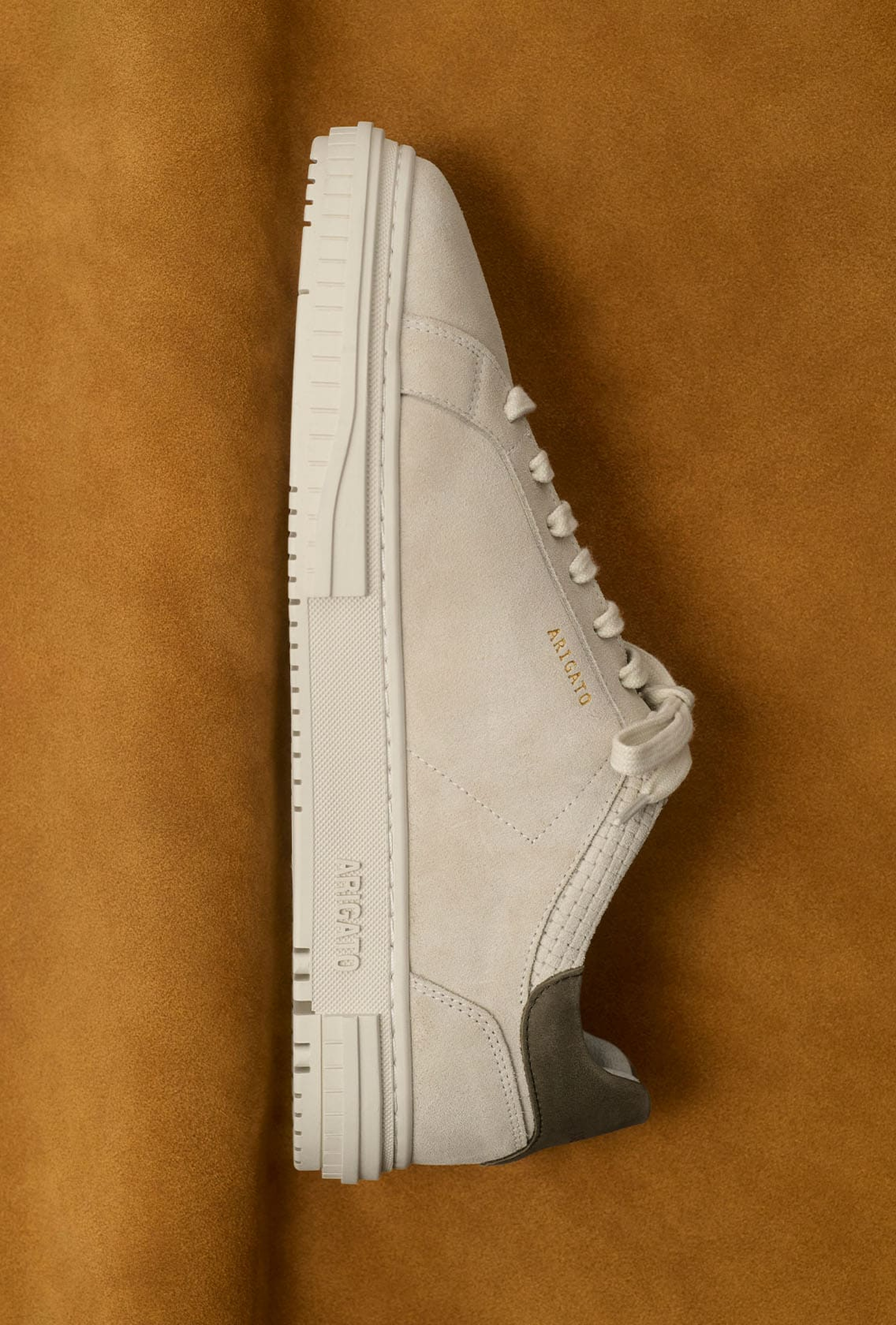 Side view of a beige suede sneaker with white sole, gold branding, and gray heel detail, on a brown background.