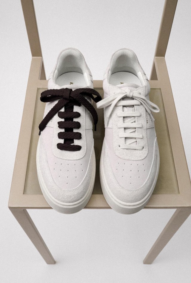 Two white sneakers on a chair; left shoe has thick black laces, right shoe has thin white laces.