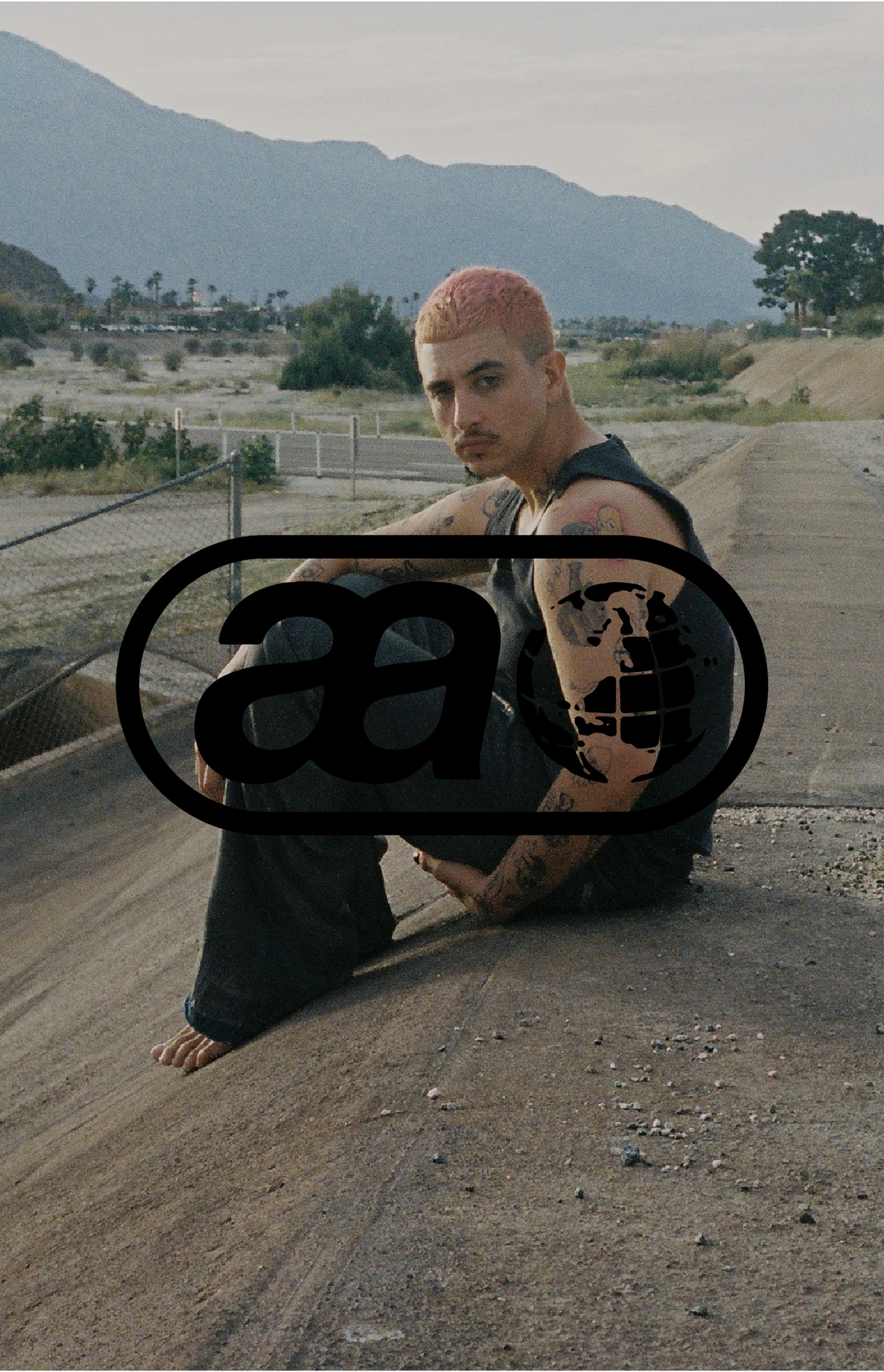 Person with short pink hair and tattoos sits on a sloped concrete surface, with mountains and trees in the background.