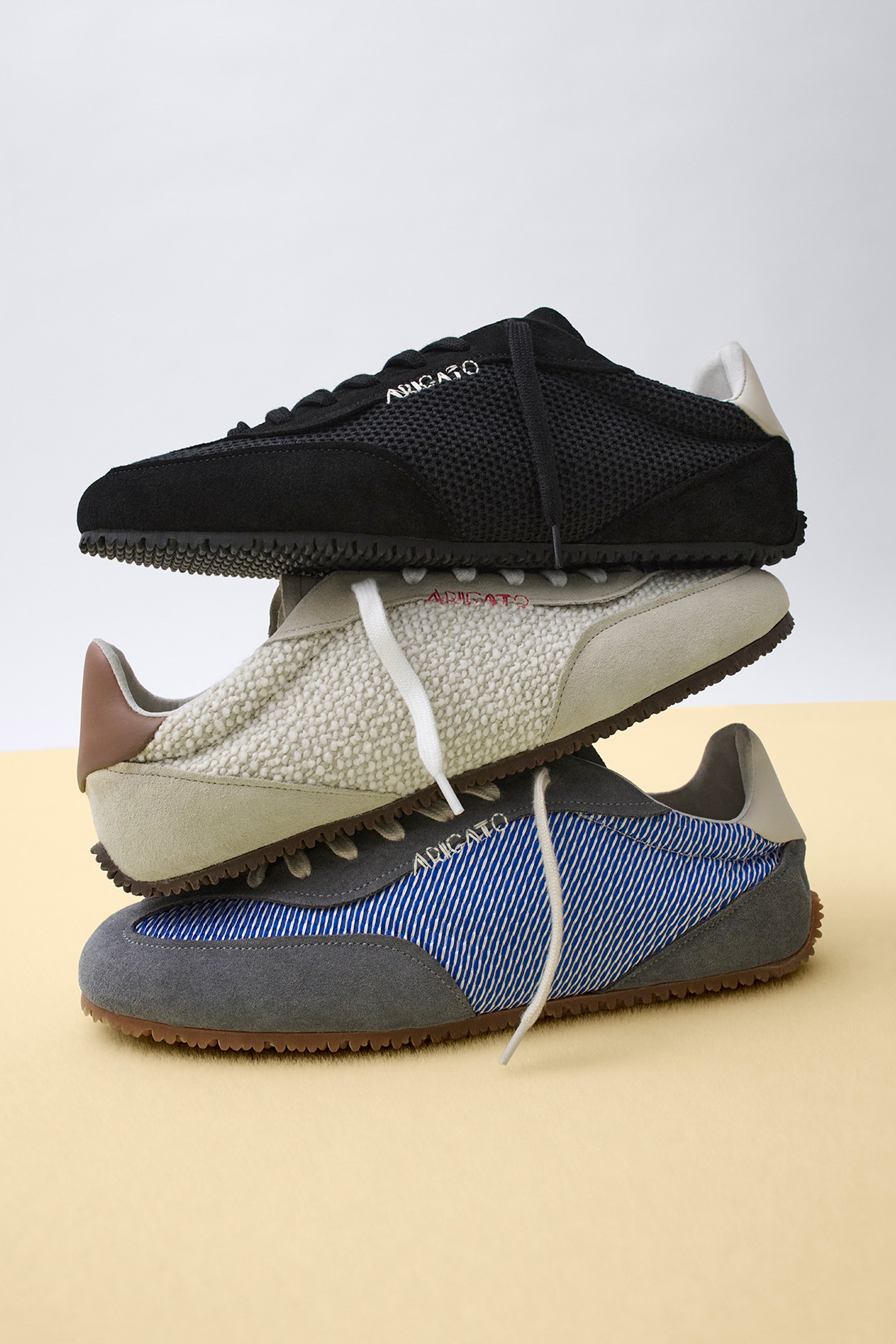 Three sneakers stacked: black mesh on top, beige textured in the middle, and blue-striped with gray suede at the bottom, on a yellow surface.