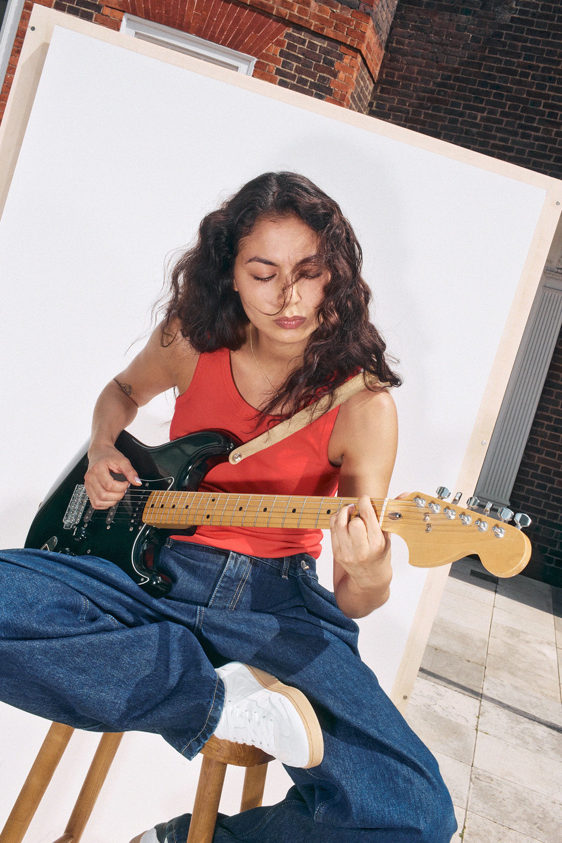 Woman in red tank top and jeans playing an electric guitar, seated on a stool against a white backdrop.