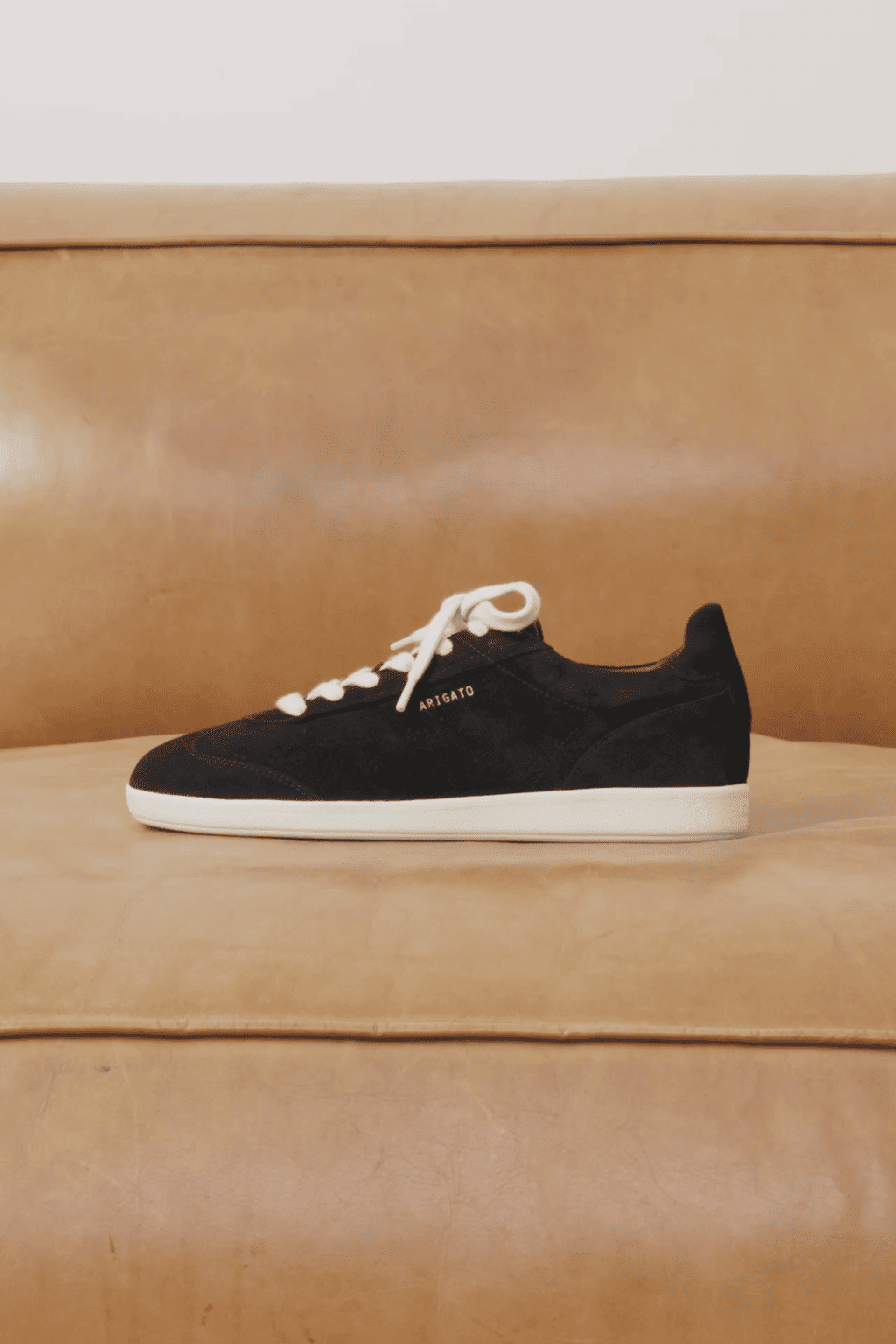 Black suede sneaker with white laces and sole, labeled "APRS ATO," displayed on a tan leather couch.