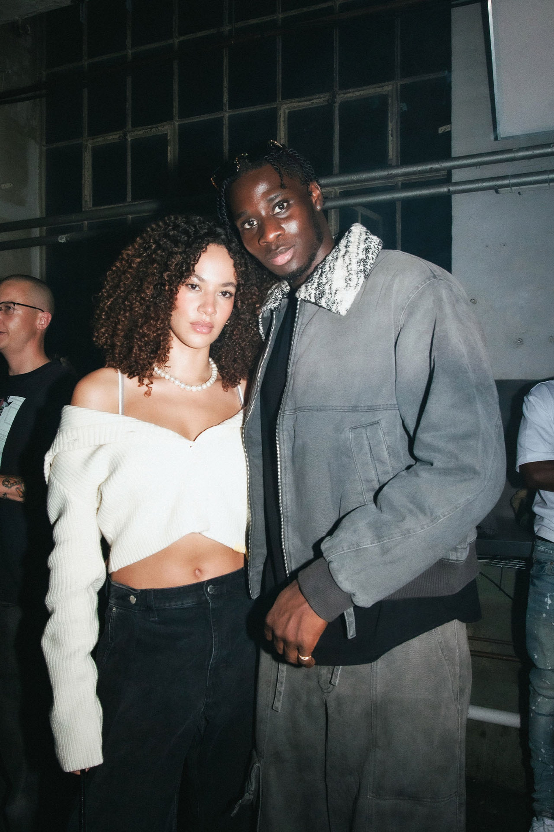 A couple poses together at an indoor event, with the woman wearing a white off-shoulder top and the man in a dark jacket.