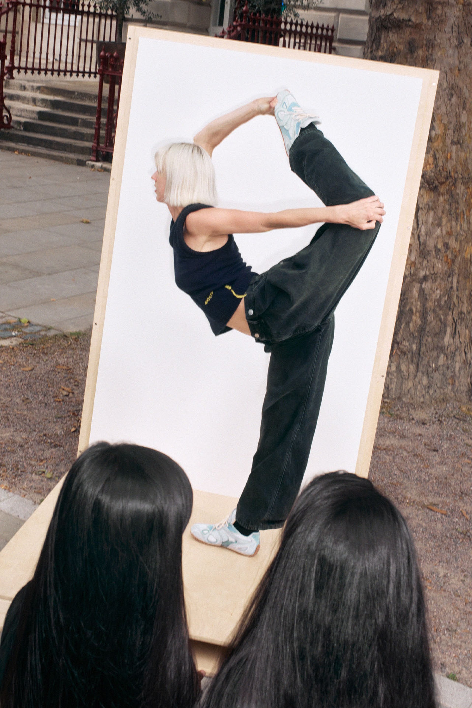 A person with blonde hair performs a standing split in front of a white backdrop, observed by two people with long dark hair.