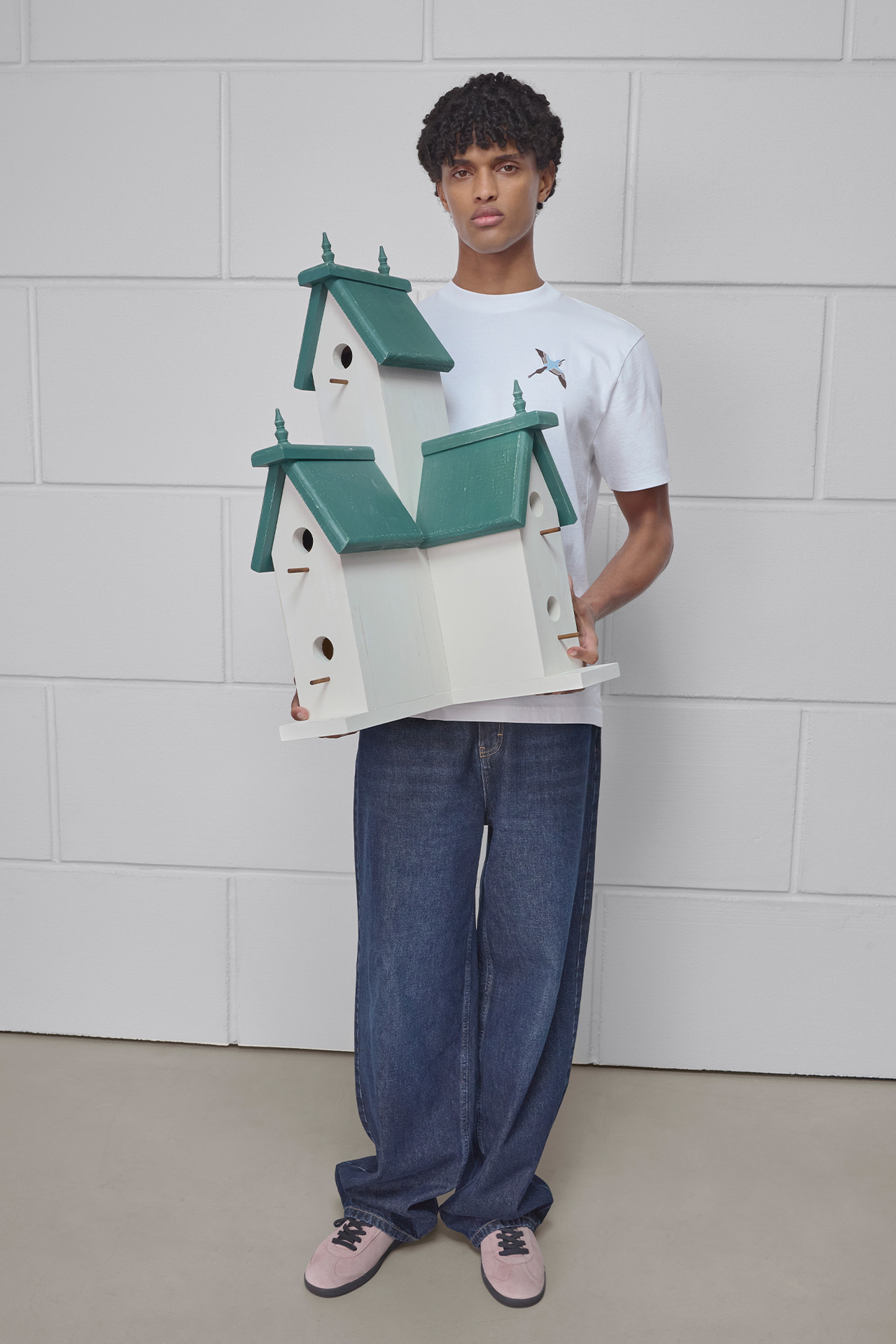 A person in a white shirt and blue jeans holds a large, multi-level birdhouse with green roofs against a plain white background.