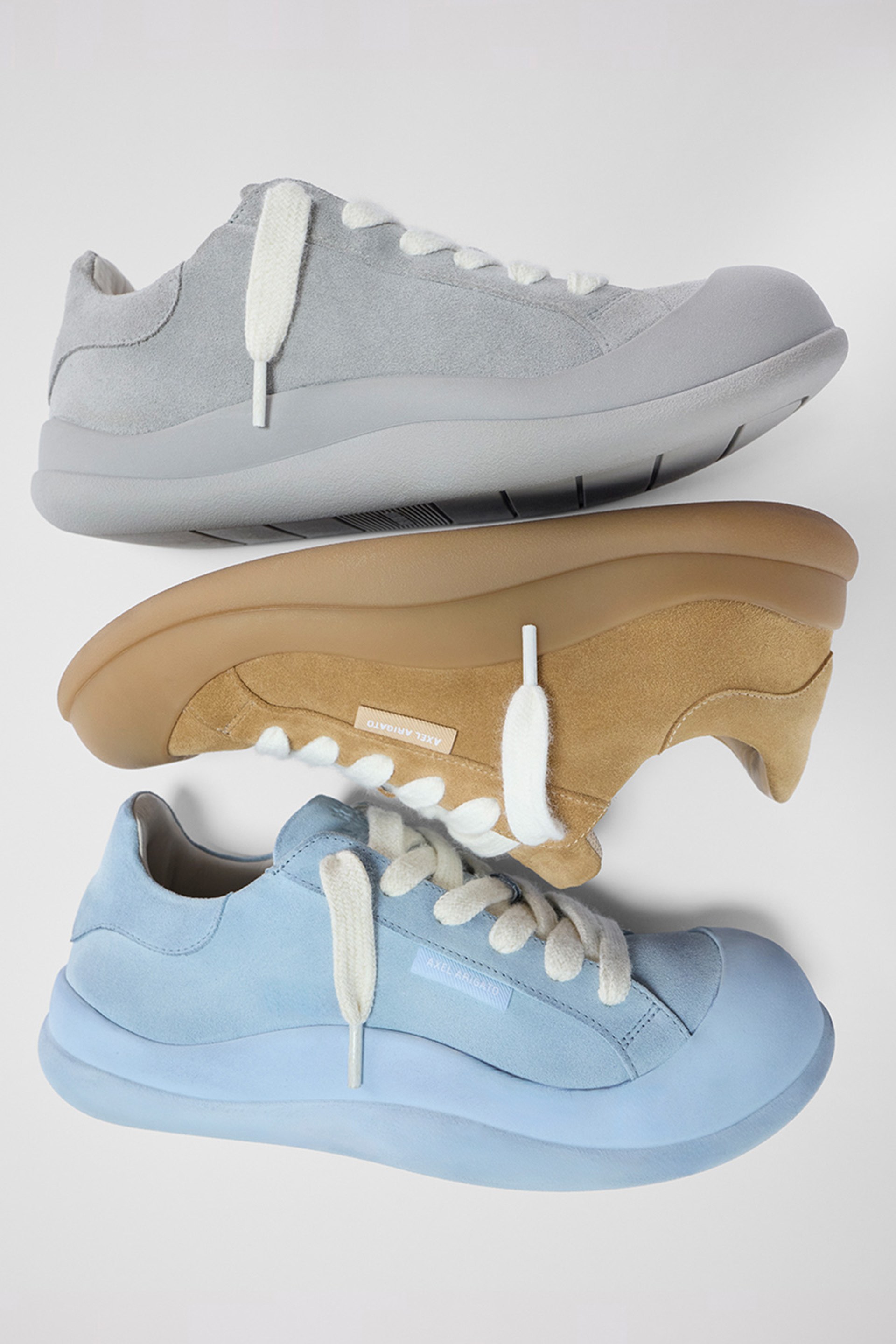 Three casual sneakers stacked; grey, tan, and blue. Each has white laces and a thick sole. Minimalist design on a white background.