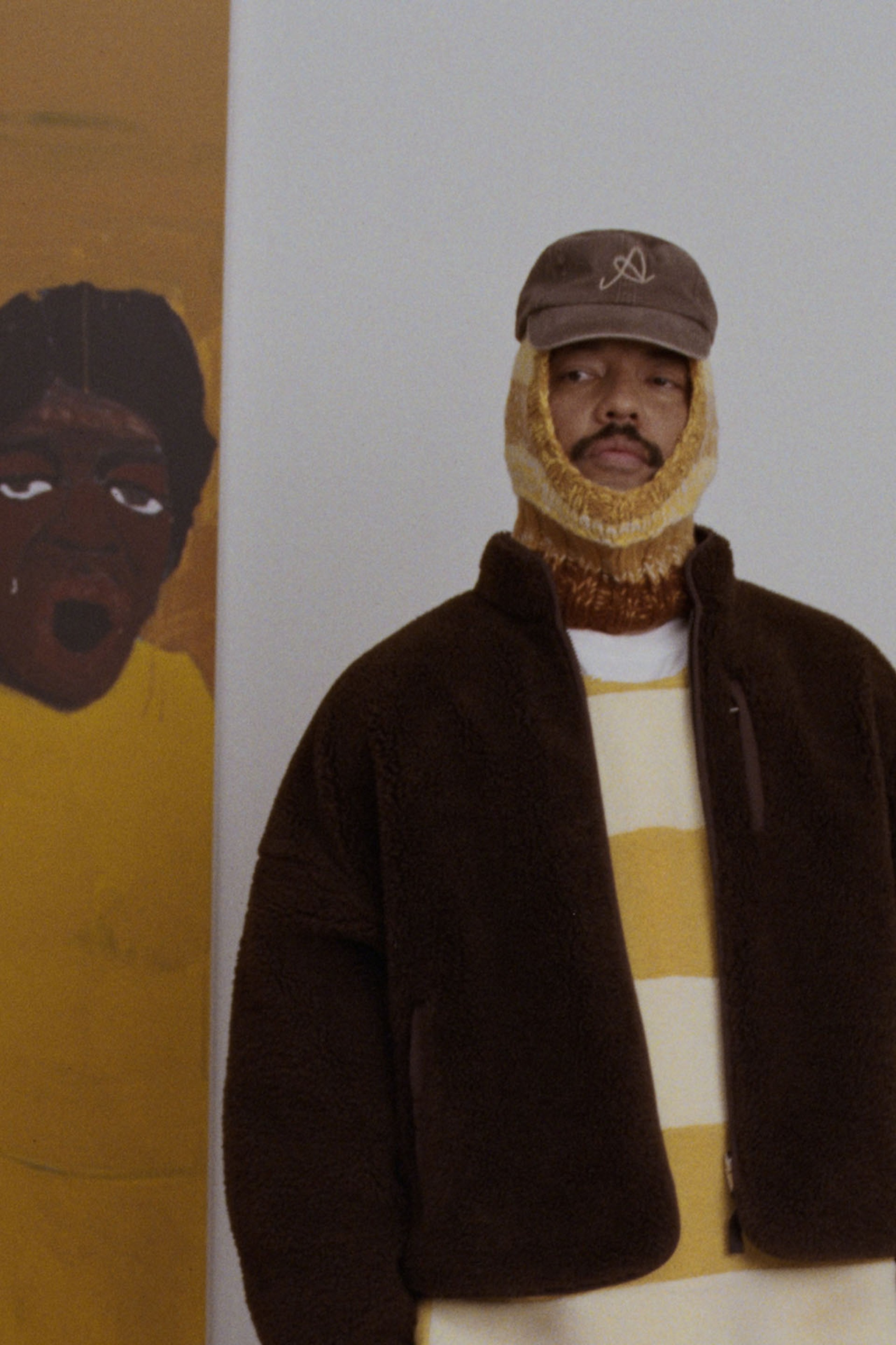 Man in a brown jacket and striped sweater, wearing a cap and knitted face covering, stands next to a painting of a person in yellow.
