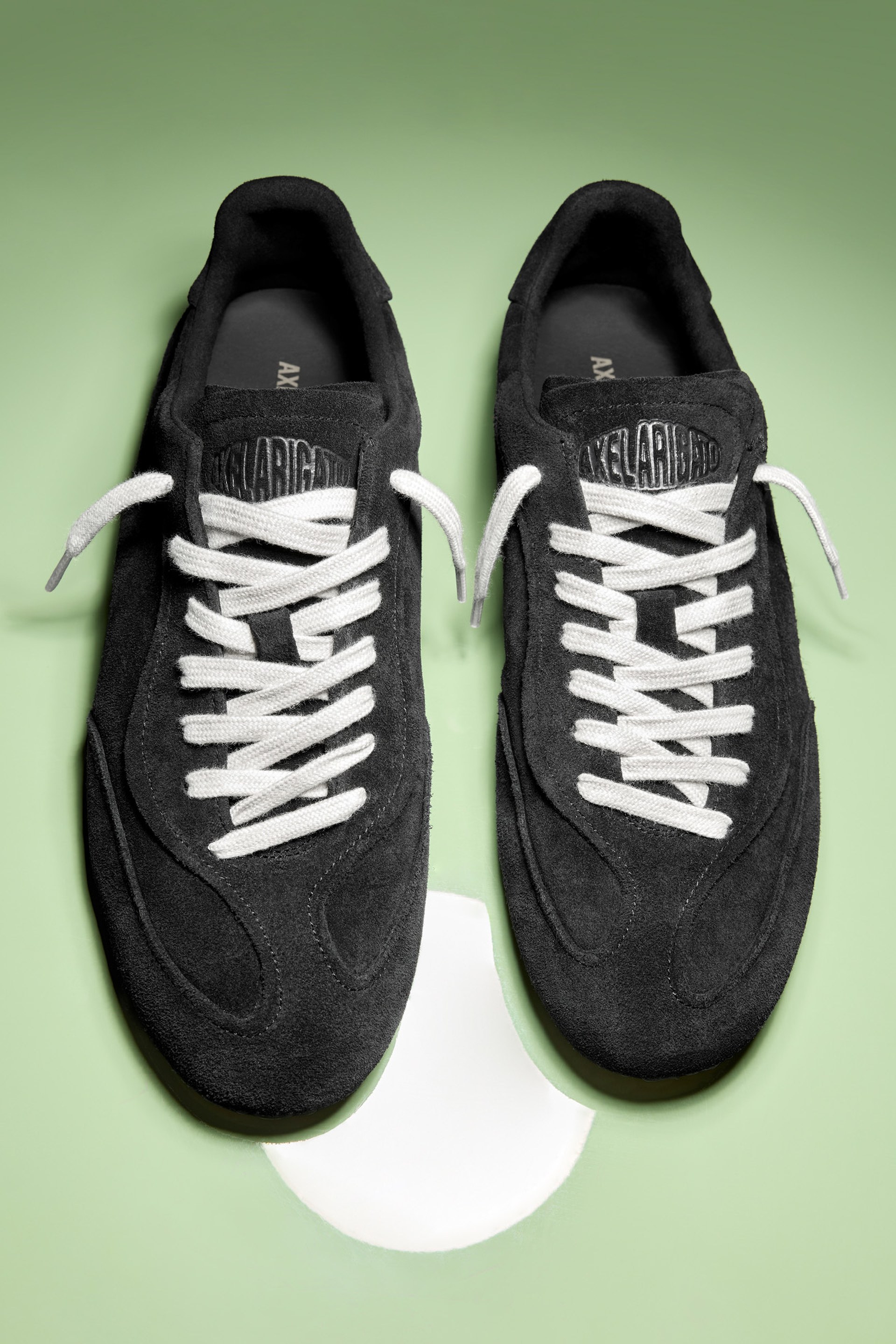 Black suede sneakers with white laces on a light green background, positioned side by side, top view.