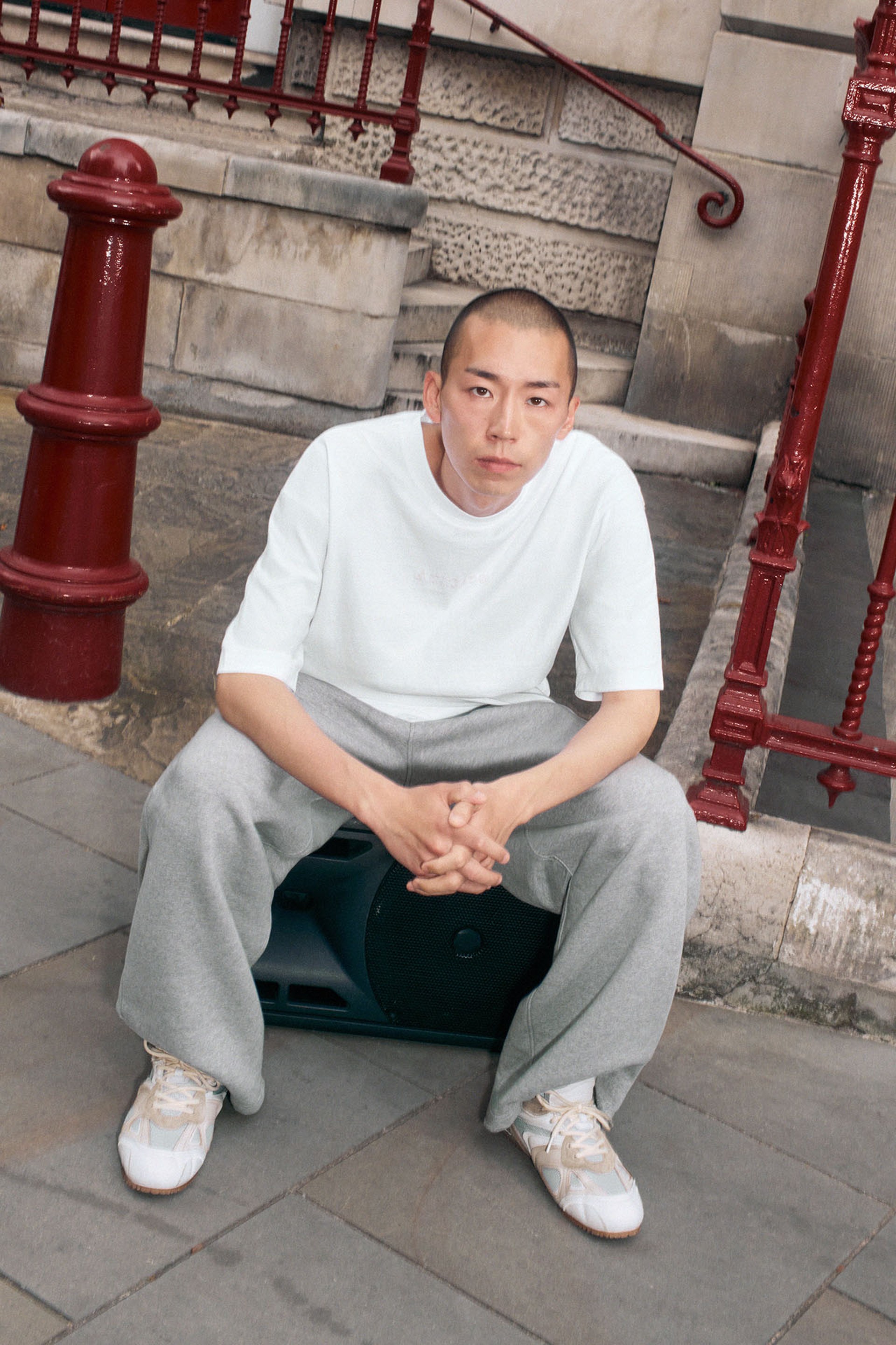 Person in a white shirt and gray pants sitting on a speaker outdoors, with red railings and stone steps in the background.