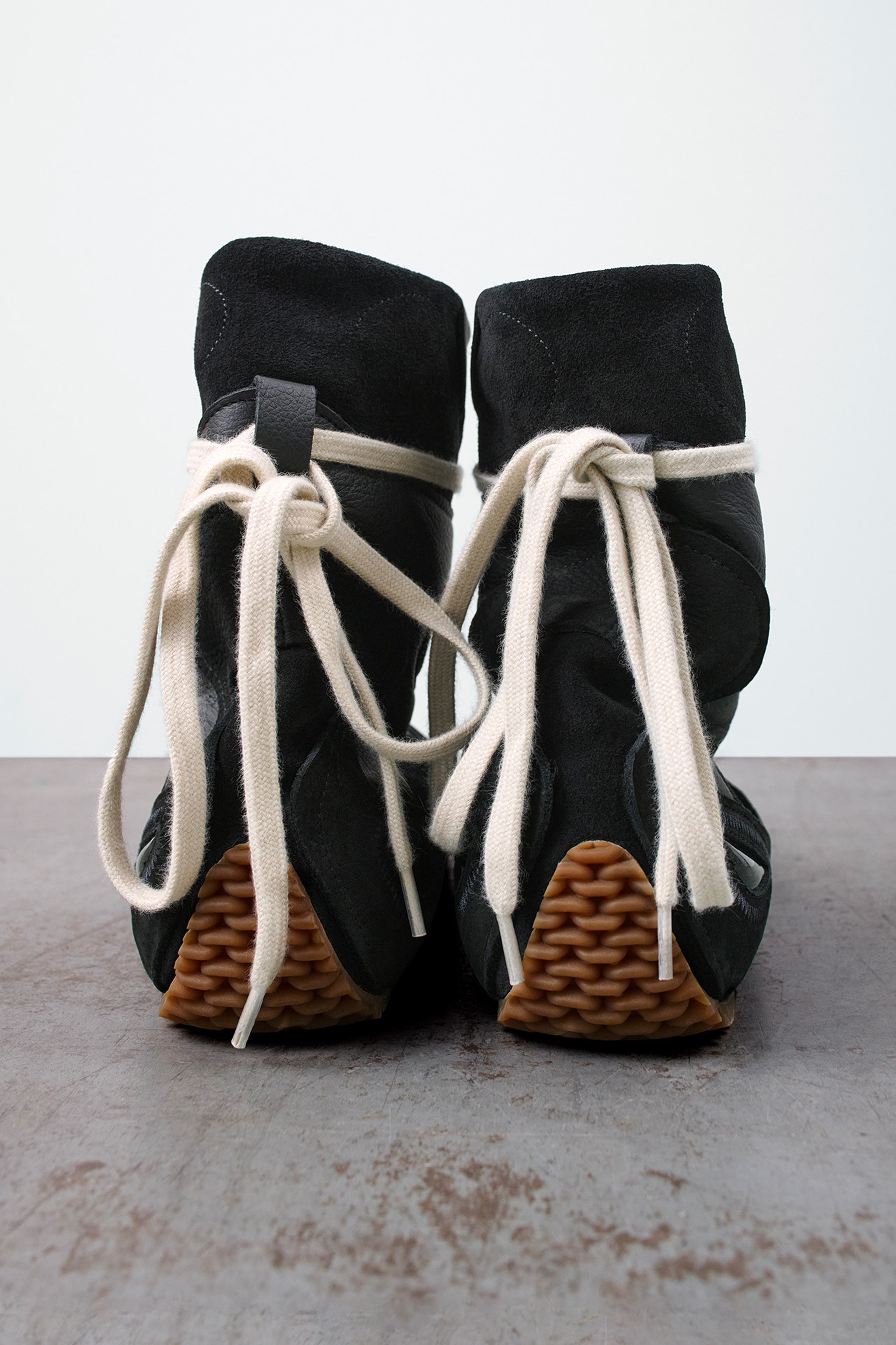 Black high-top sneakers with white laces, viewed from the back, featuring textured brown soles on a gray surface.