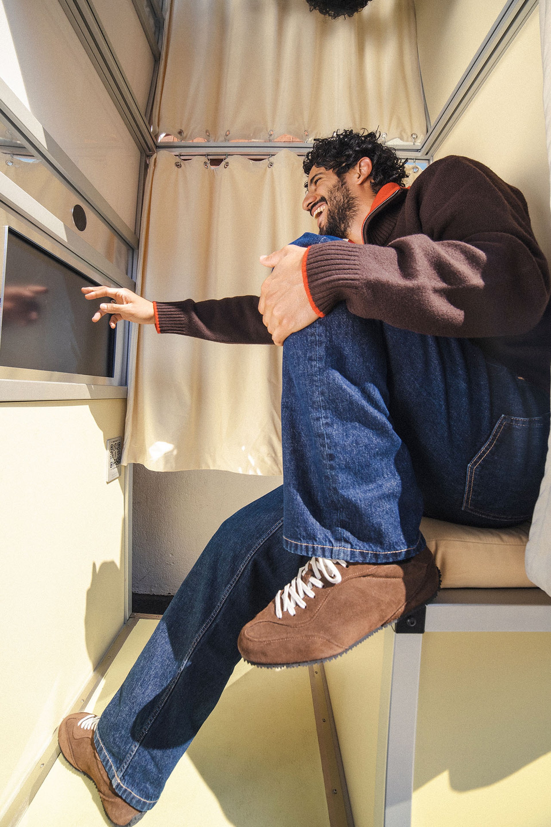 A man in a cozy, compact space laughs while reaching towards a screen. He sits with his leg up, wearing a brown jacket and jeans.