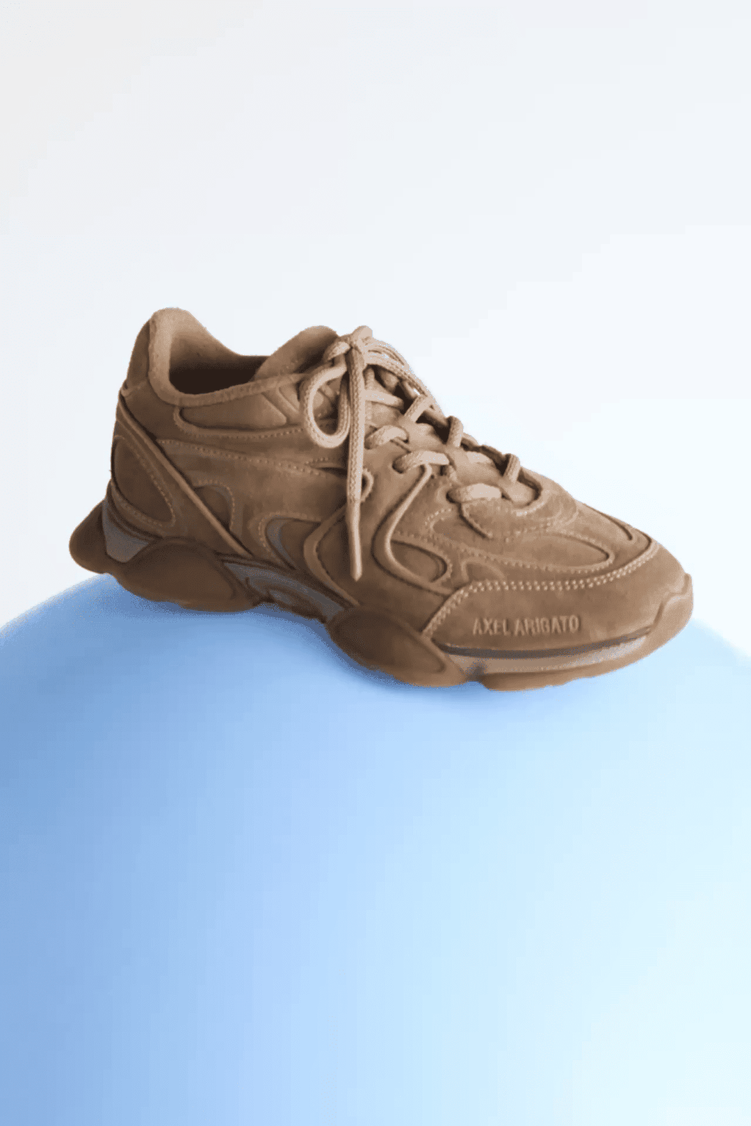Brown sneaker with intricate stitching and "AXEL ARIGATO" text, placed on a light blue surface against a white background.