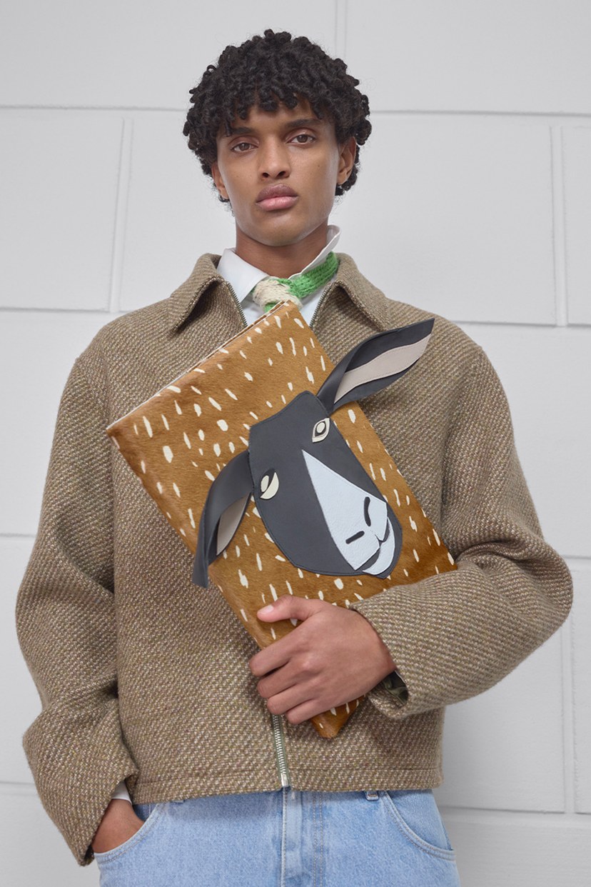 Person in a brown jacket holds a rectangular bag with an illustrated animal face on it, standing against a white tiled wall.