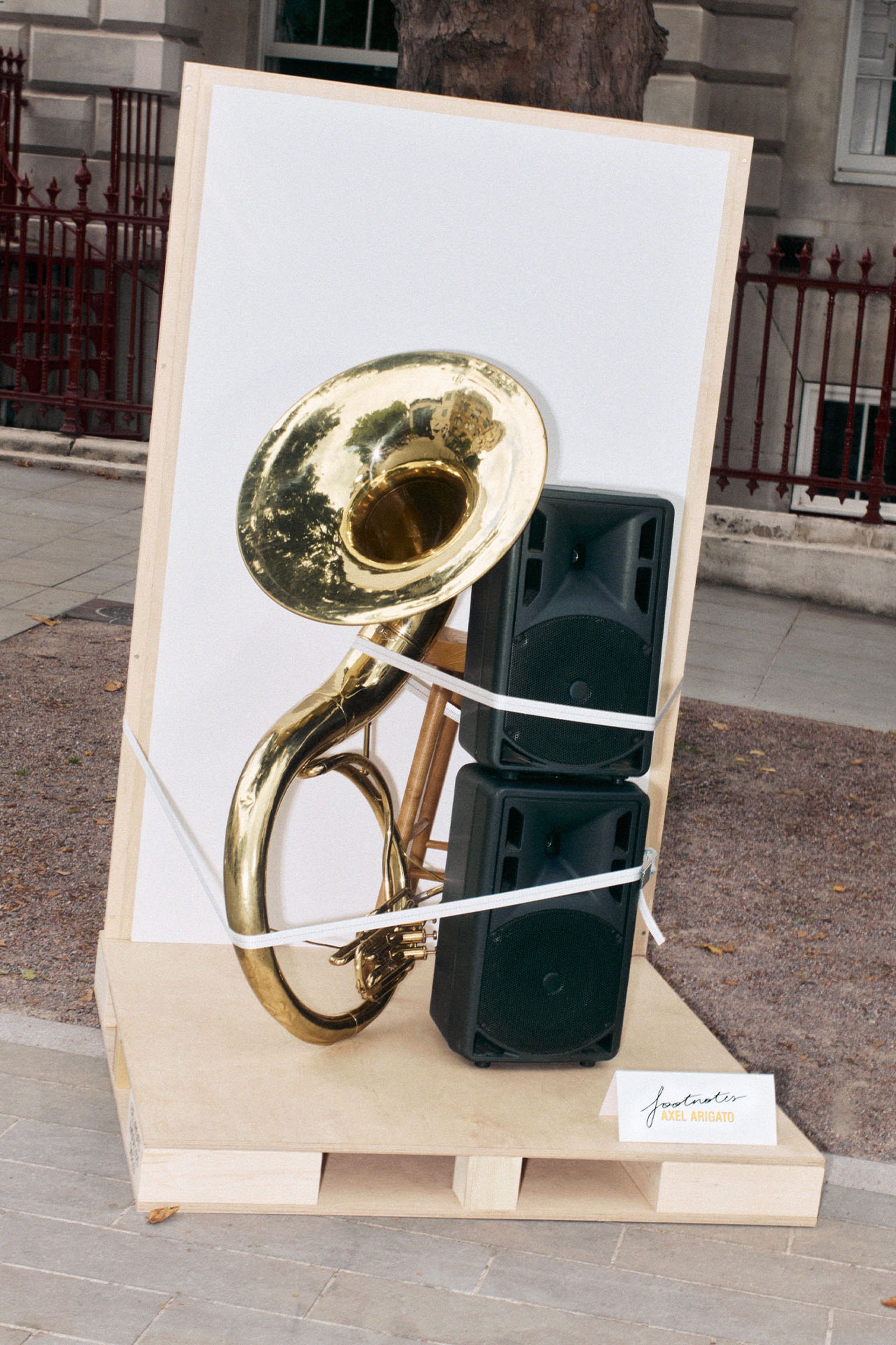 A sousaphone strapped to two large speakers, displayed on a wooden platform outdoors, with a small sign nearby.