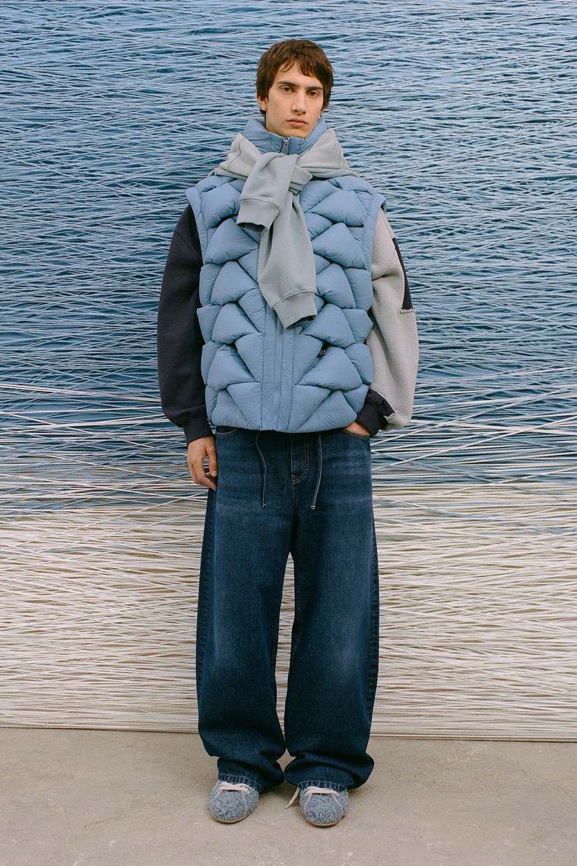 Person wearing a blue puffer vest, scarf, wide-leg jeans, and gray shoes stands against a backdrop of water and sand.