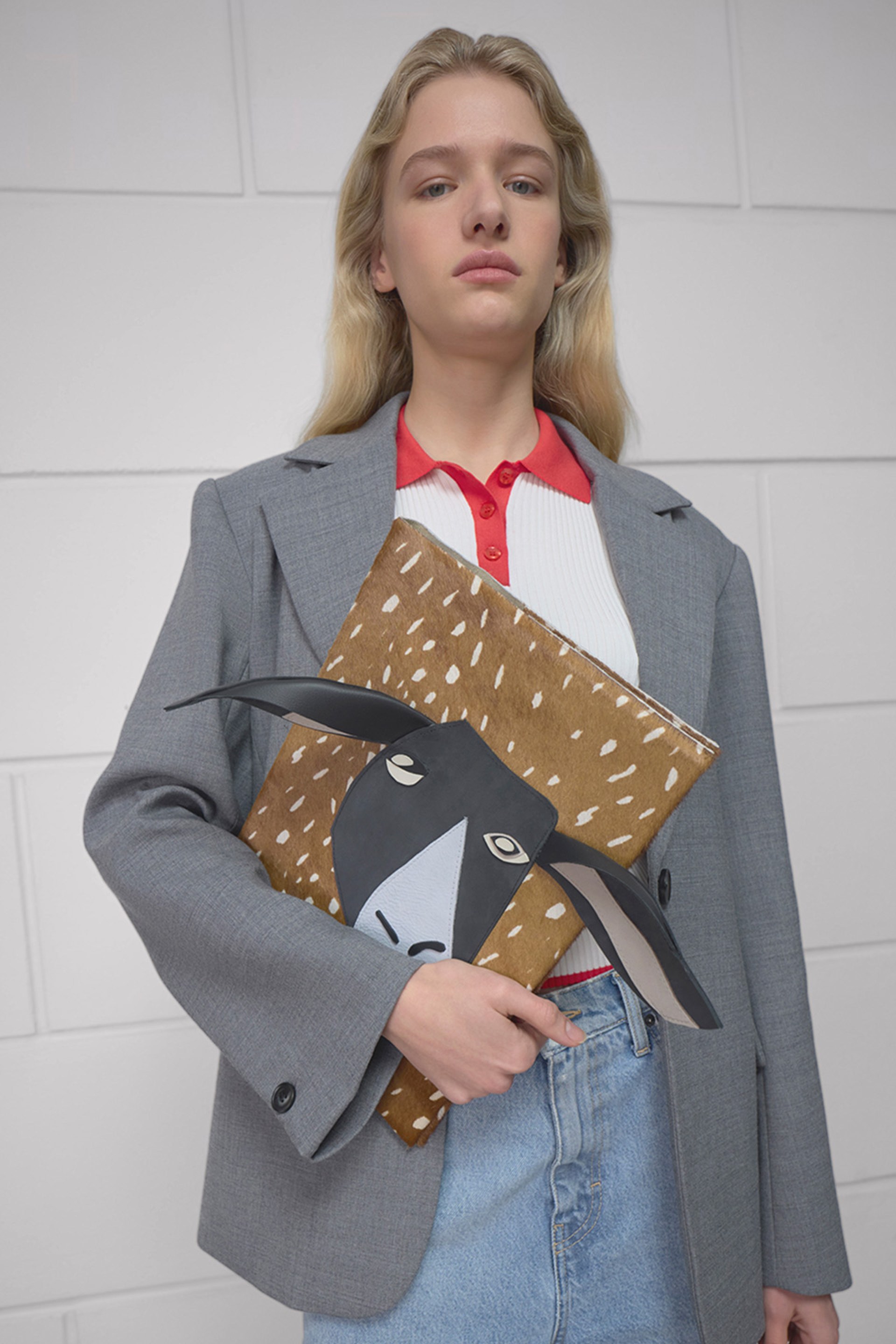 A person in a gray blazer holds a book with an animal illustration, wearing a red and white shirt and jeans, standing against a white tiled wall.