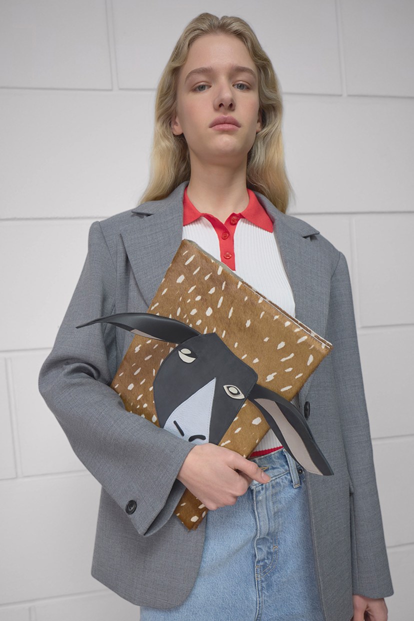 A person in a gray blazer holds a book with an animal illustration, wearing a red and white shirt and jeans, standing against a white tiled wall.