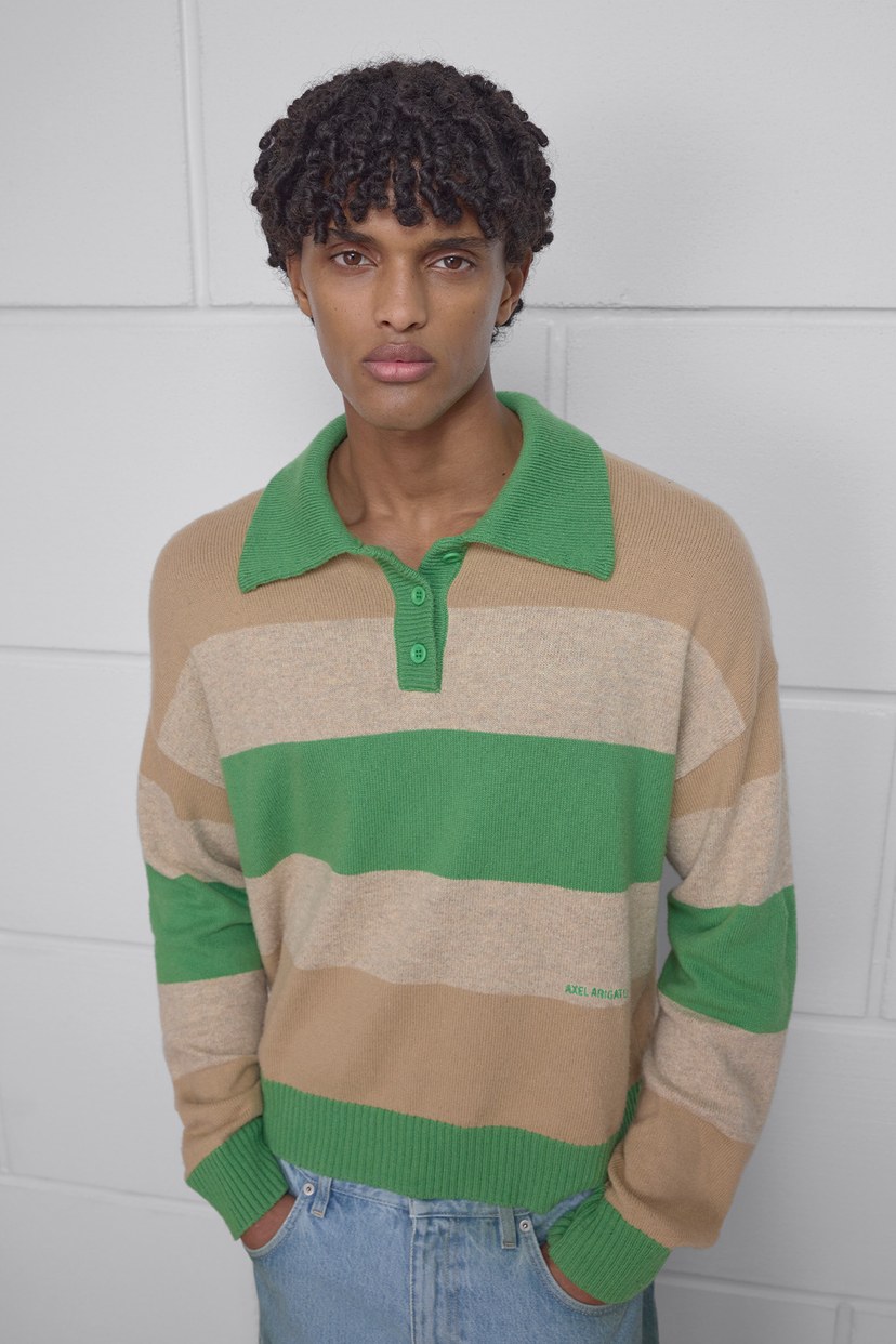 A person with curly hair wears a green and beige striped sweater and light blue jeans, standing against a white wall.