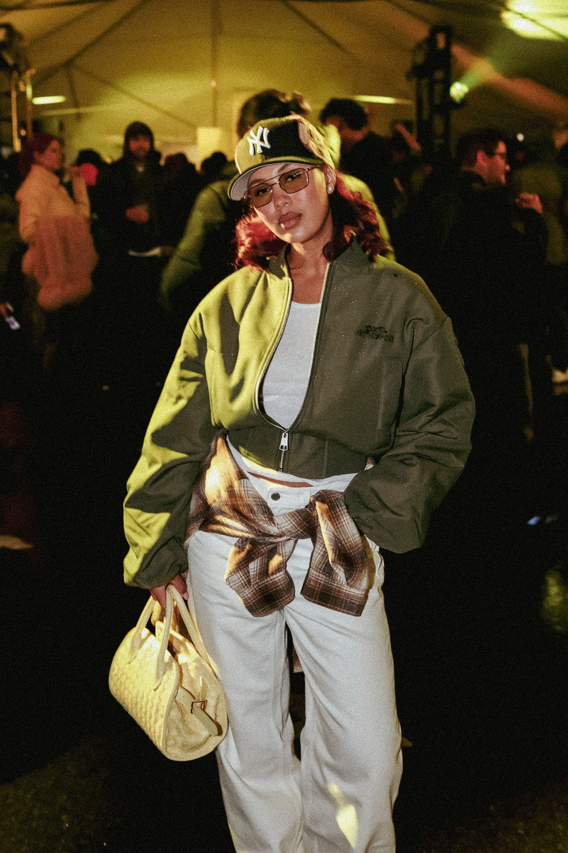 Woman in a green jacket and cap, wearing glasses, carries a woven bag at a dimly lit event with people mingling in the background.