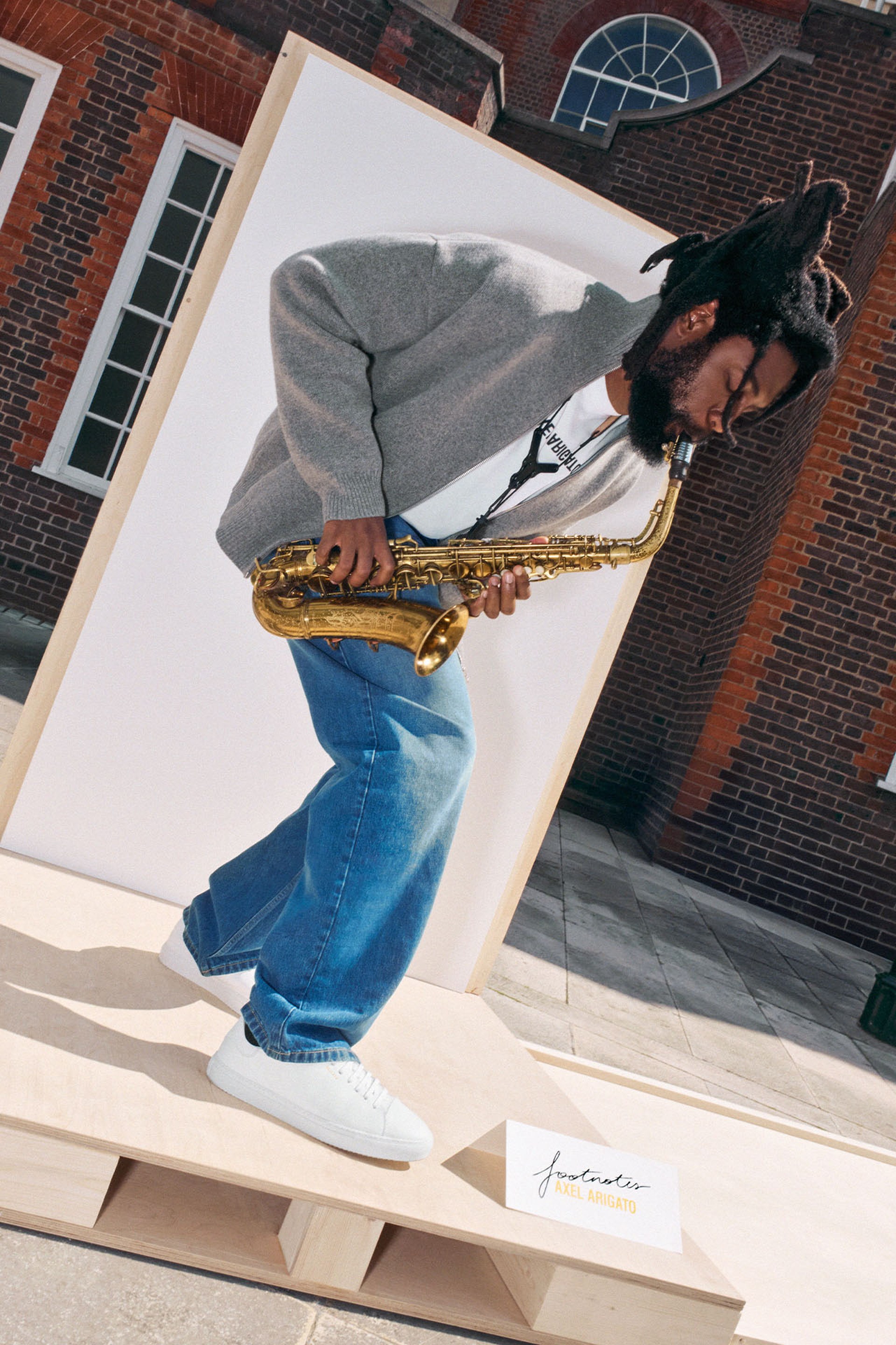 Person playing a saxophone, wearing a gray jacket and jeans, stands on a wooden platform against a brick building backdrop.
