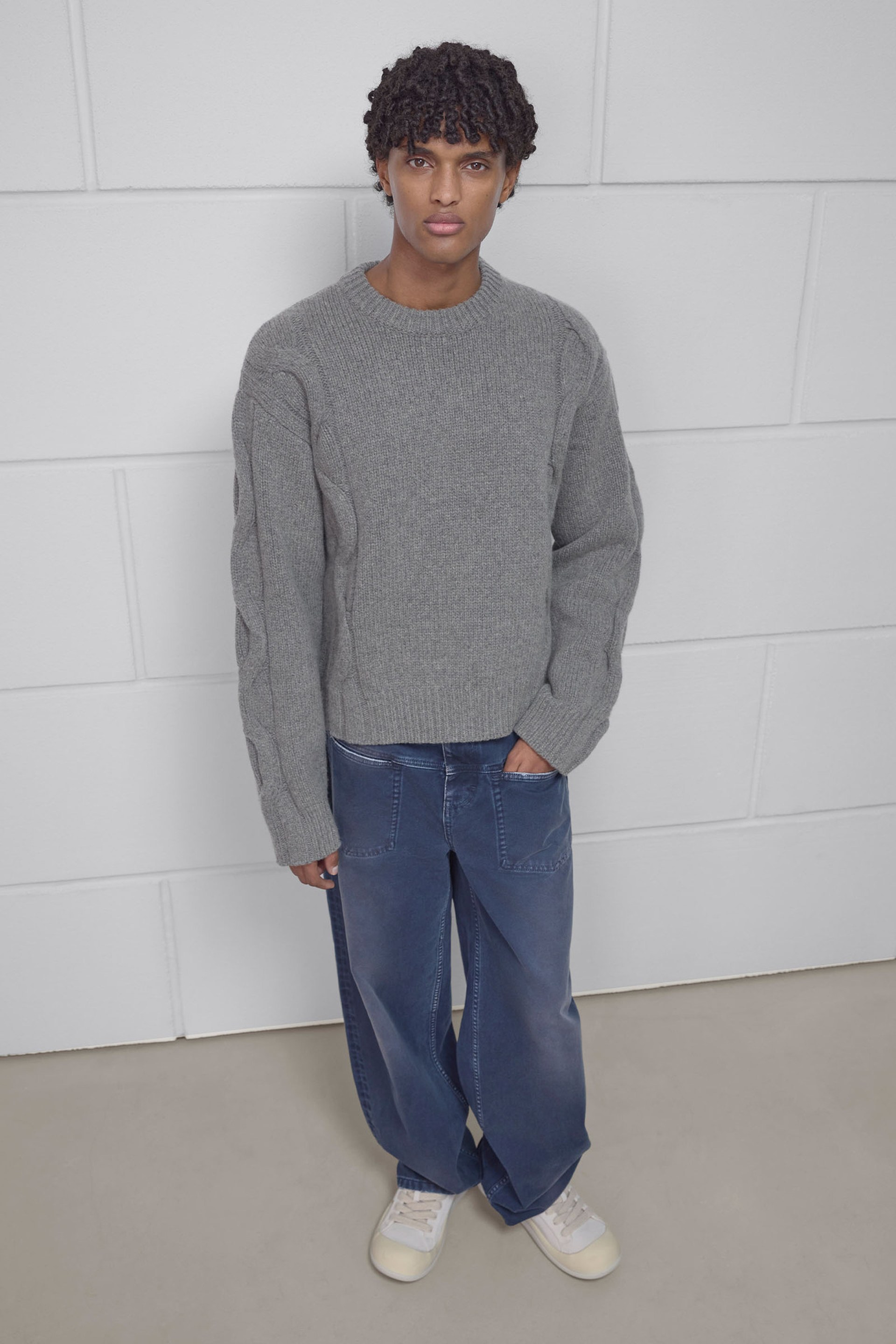 Person wearing a gray sweater and blue wide-legged jeans, standing against a white brick wall, with hands in pockets.