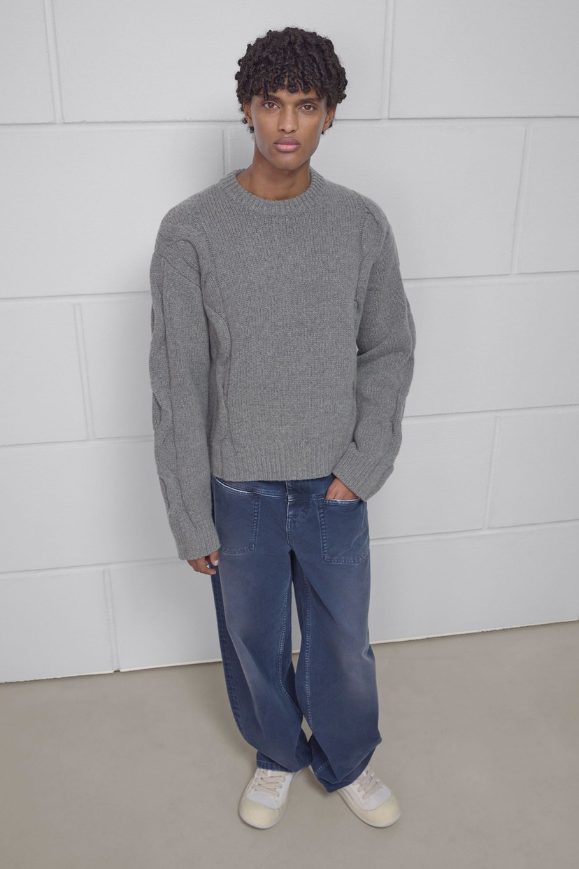 A person with curly hair wearing a gray sweater and blue wide-leg jeans stands against a white tiled wall, hands in pockets.