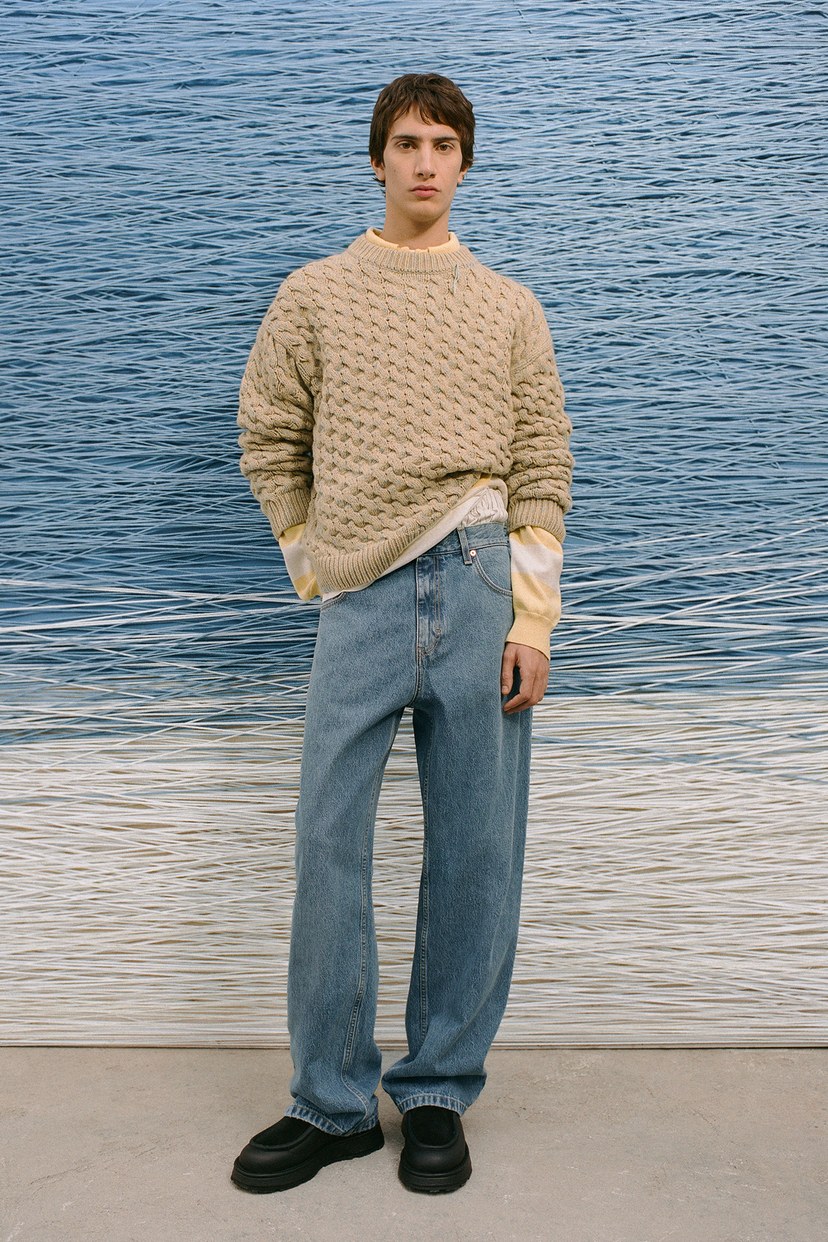 Person in a beige knit sweater and blue jeans stands against a textured blue and white background.