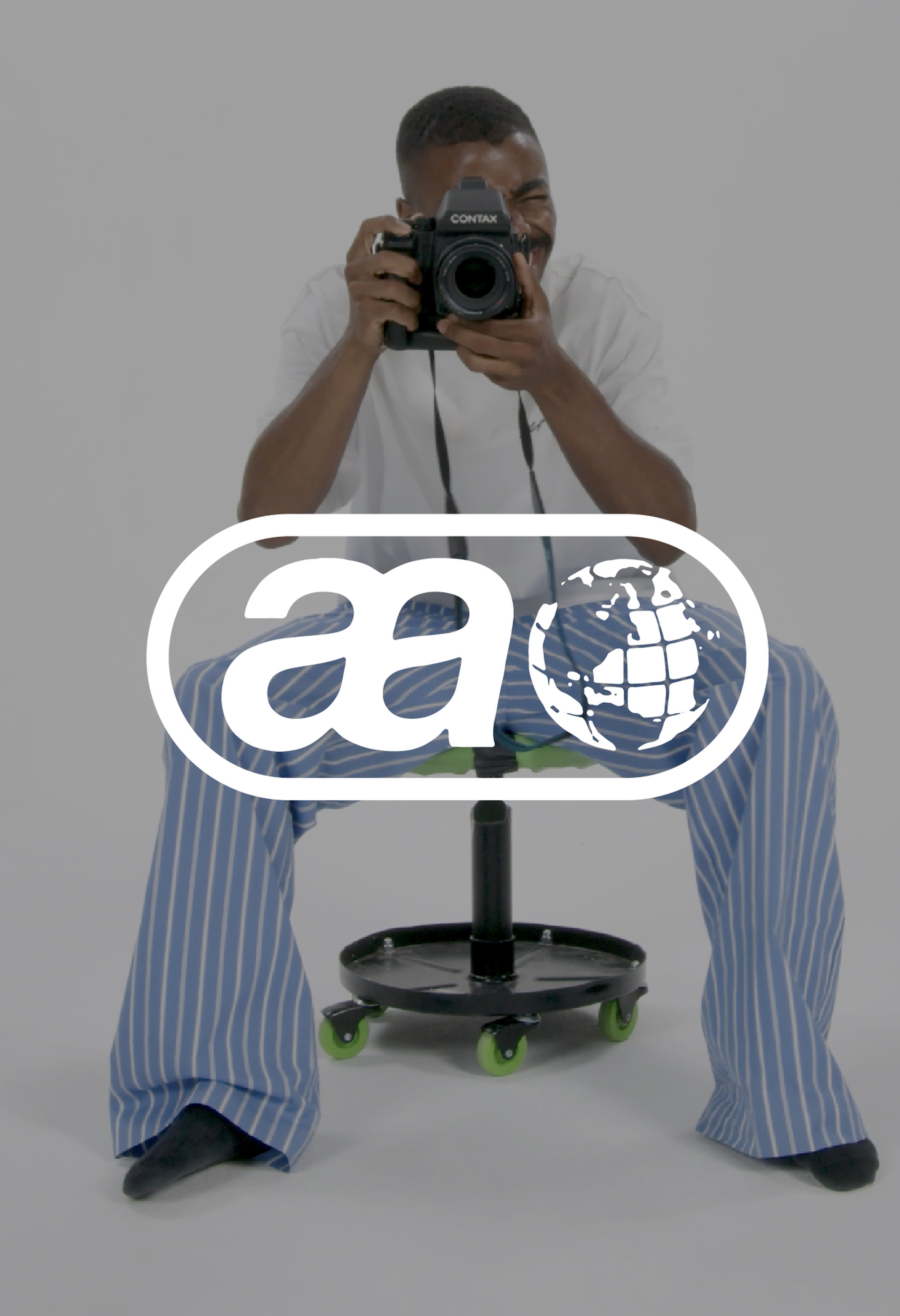 Person sitting on a stool, holding a camera to their face, wearing a white shirt and blue-striped pants against a plain background.