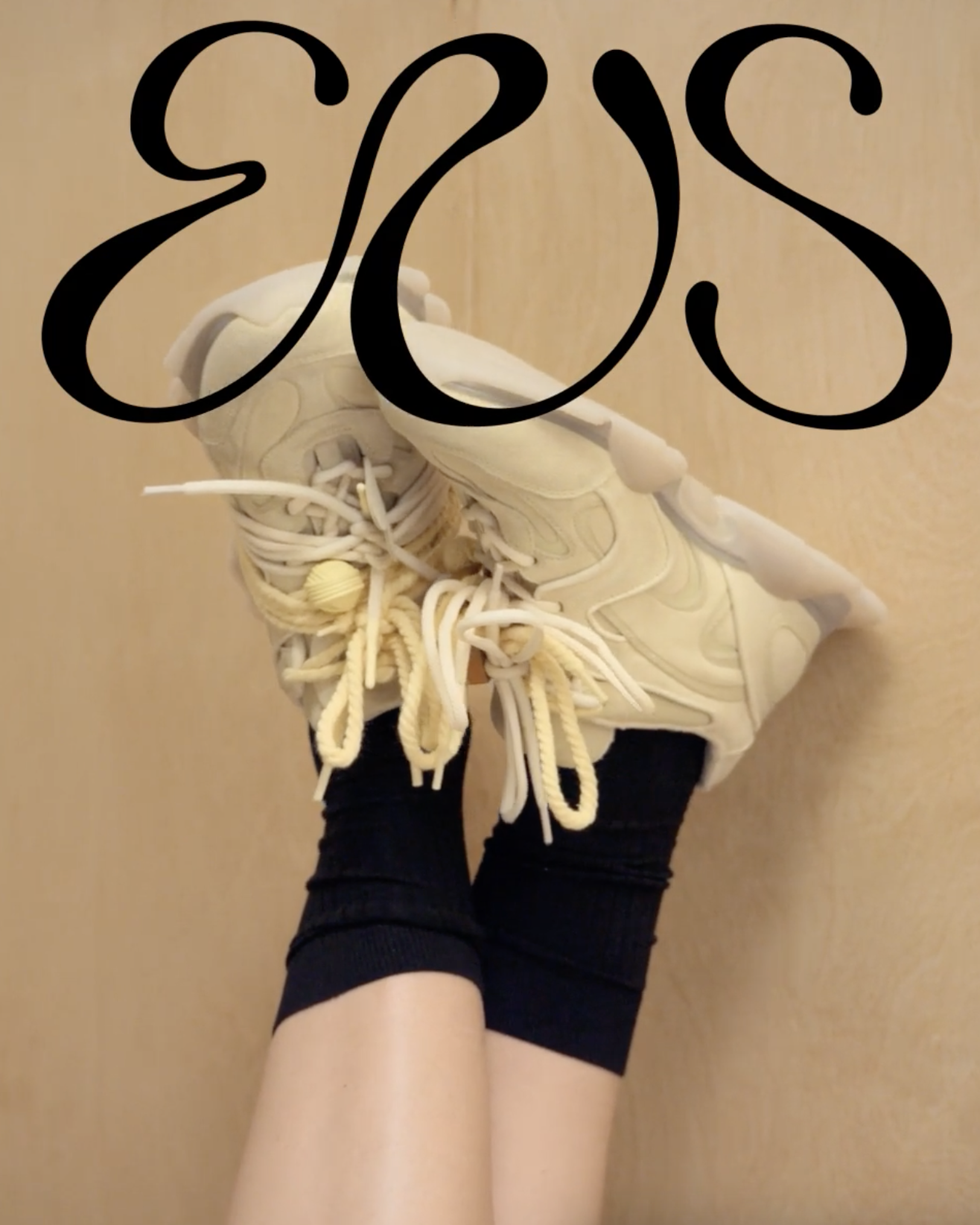 Feet wearing yellow Eris sneakers with yellow laces and black socks, raised against a wooden background. The word "EUS" is overlaid at the top.