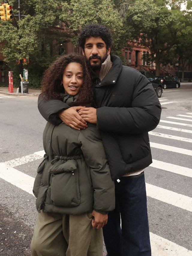 Couple standing together on a city street, both wearing winter coats, with trees and buildings in the background.