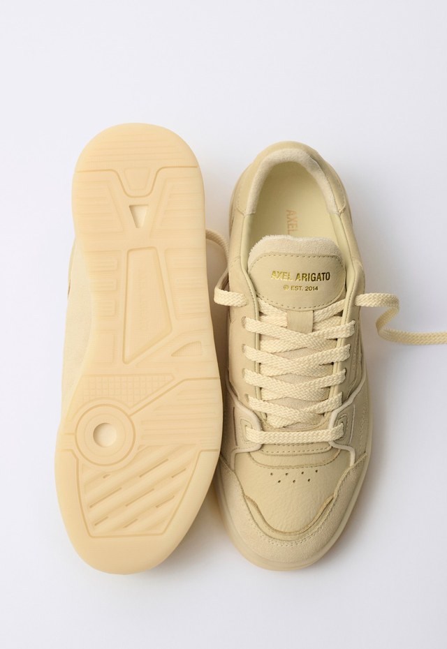 Beige sneakers with textured soles, one shoe upright showing laces, the other lying sideways displaying the sole pattern on a white background.